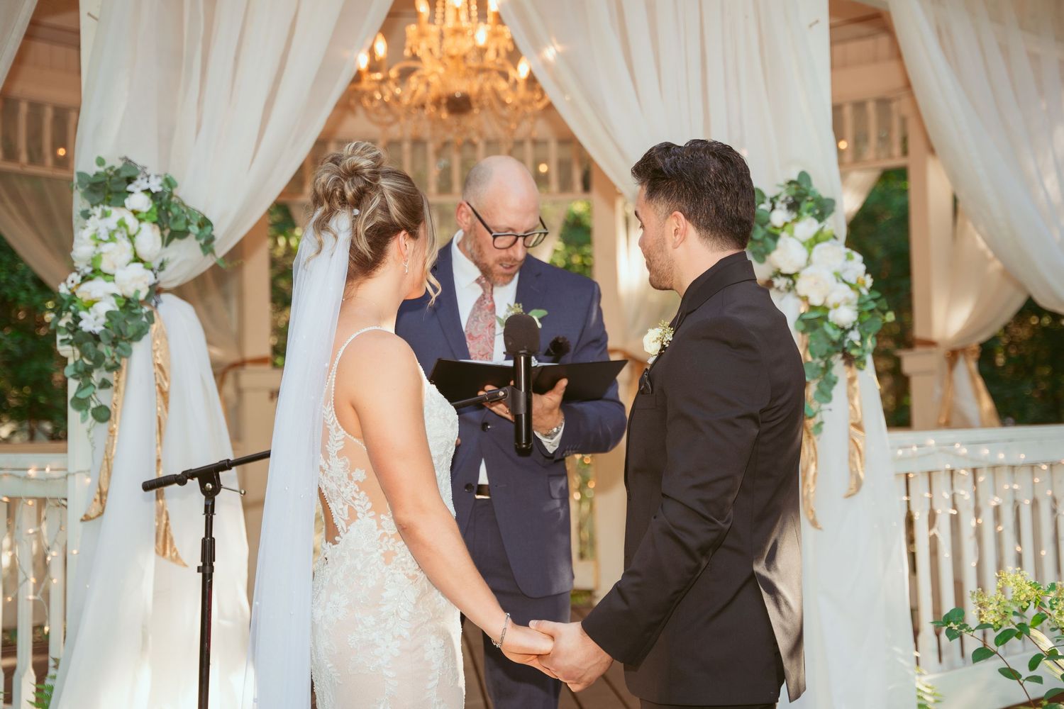 Elegant wedding ceremony setup with white drapes, chandeliers, and floral arrangements under a covered pavilion.