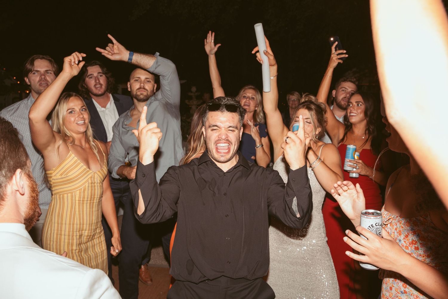 A lively wedding reception dance party with guests celebrating and raising their hands in excitement.