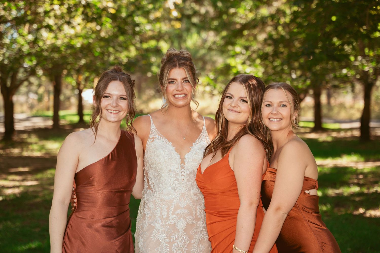 Bridal party in rust and terracotta colored dresses poses together under trees.
