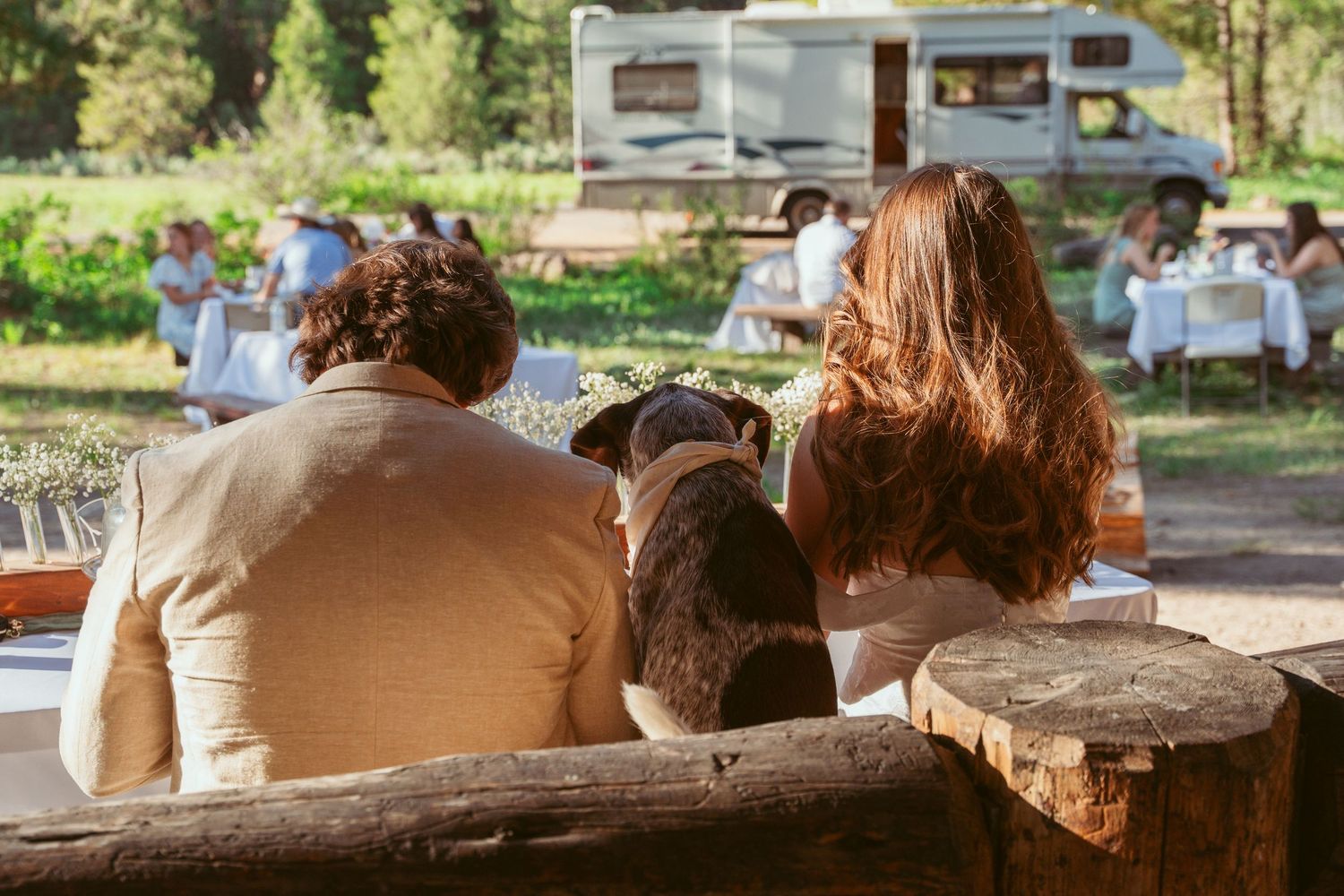 Guests relaxing on rustic log bench with RV visible in forest background.