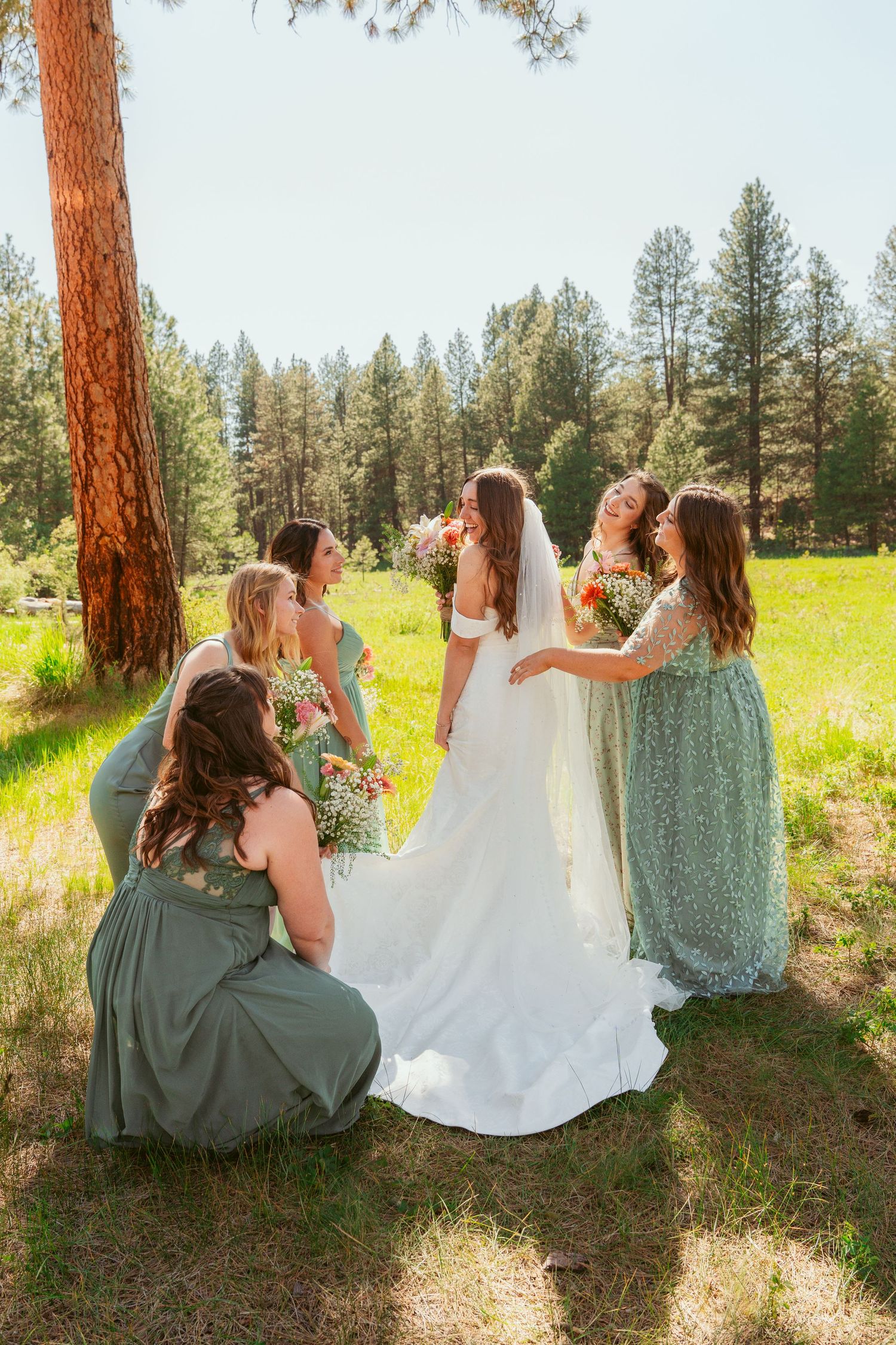 Bridesmaids in sage green dresses and bride in white gown pose together in sunny forest meadow.