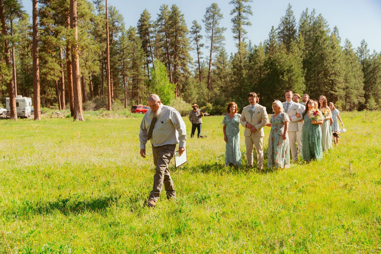 Wedding guests gather on a grassy hillside surrounded by tall pine trees for an outdoor ceremony.