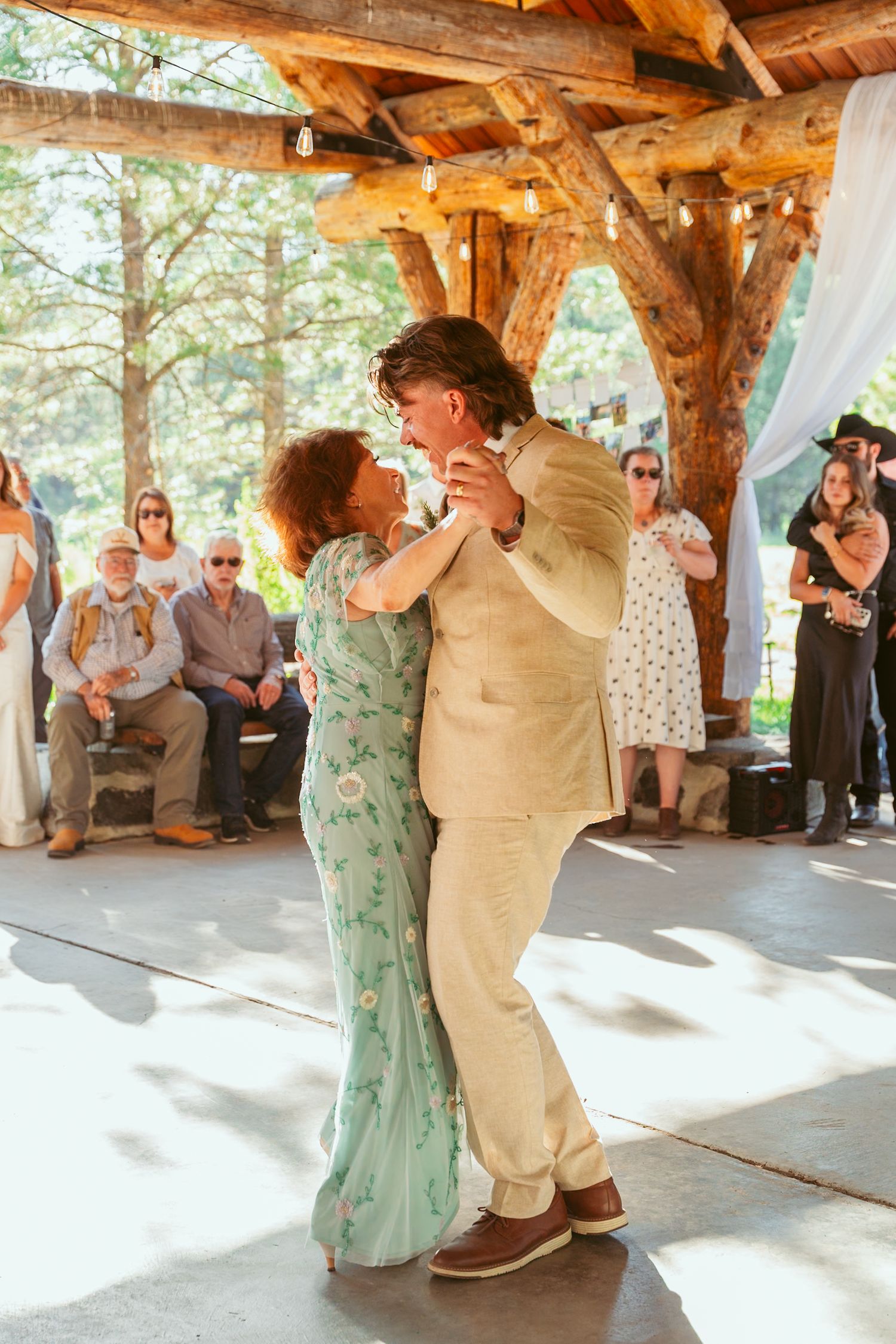 Couples dancing at outdoor wedding venue with wooden beams and forest scenery.