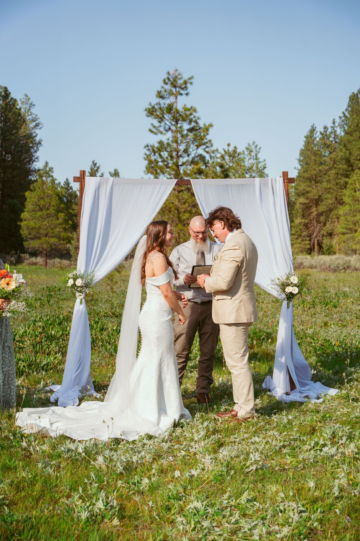 Intimate wedding ceremony moment under white fabric arch in mountain wildflower field.
