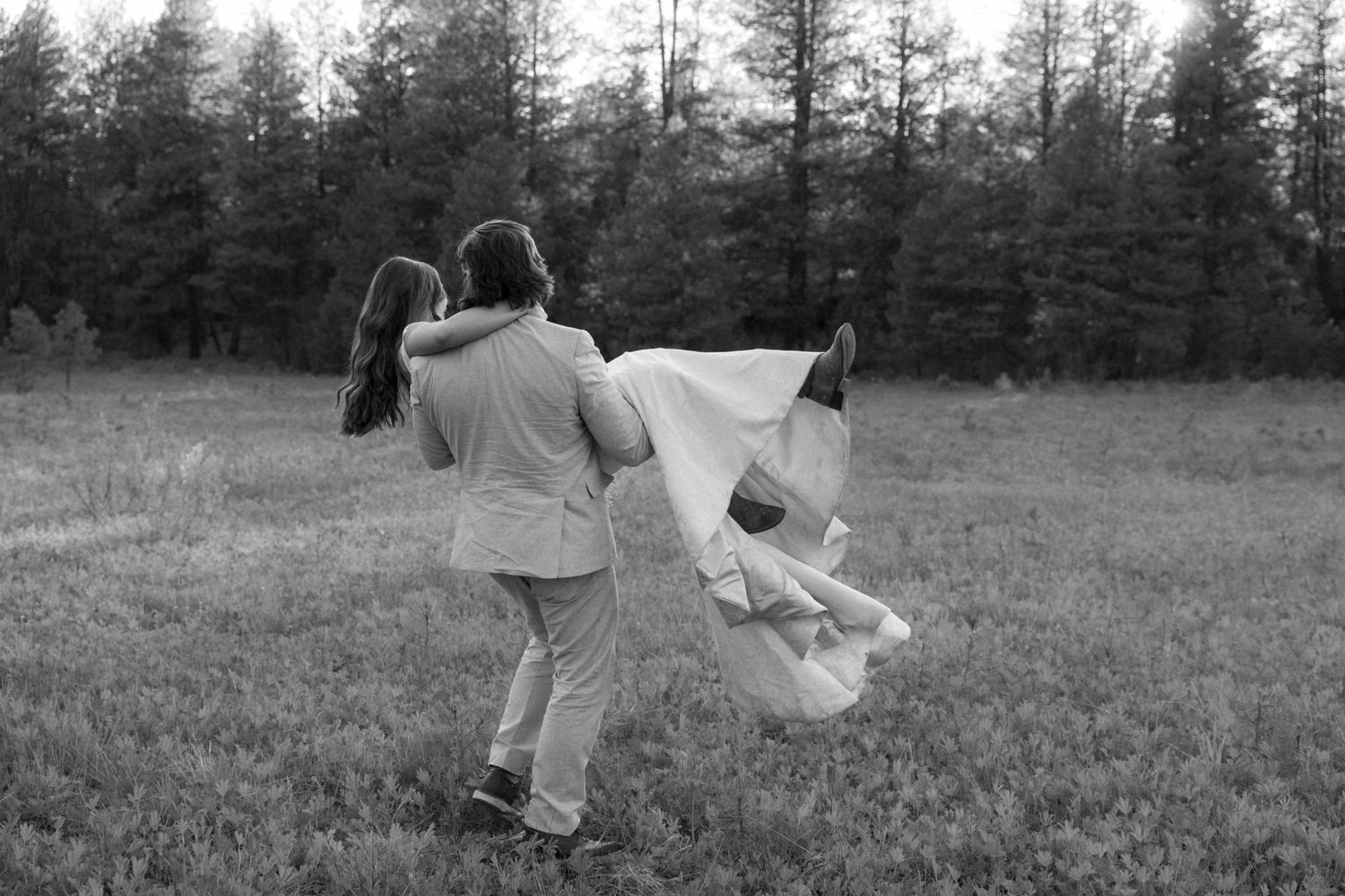 Black and white photo of a couple dancing joyfully in a field with forest backdrop.