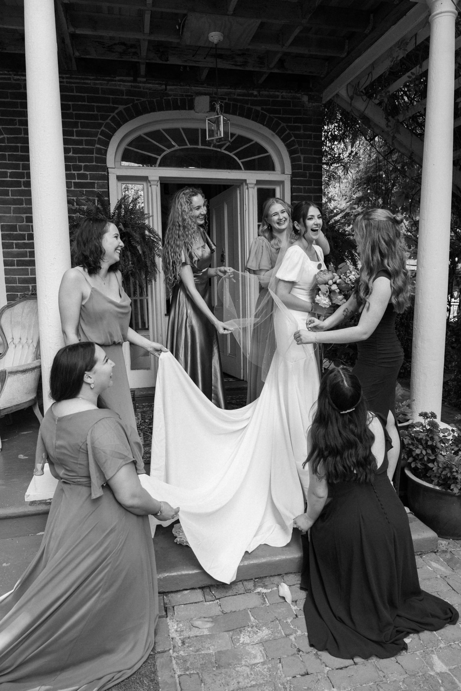 Wedding party helping bride with dress preparations on a brick porch with white columns.