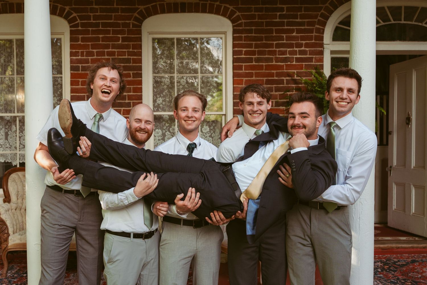 Wedding party members in gray suits act silly while posing together on columned porch.