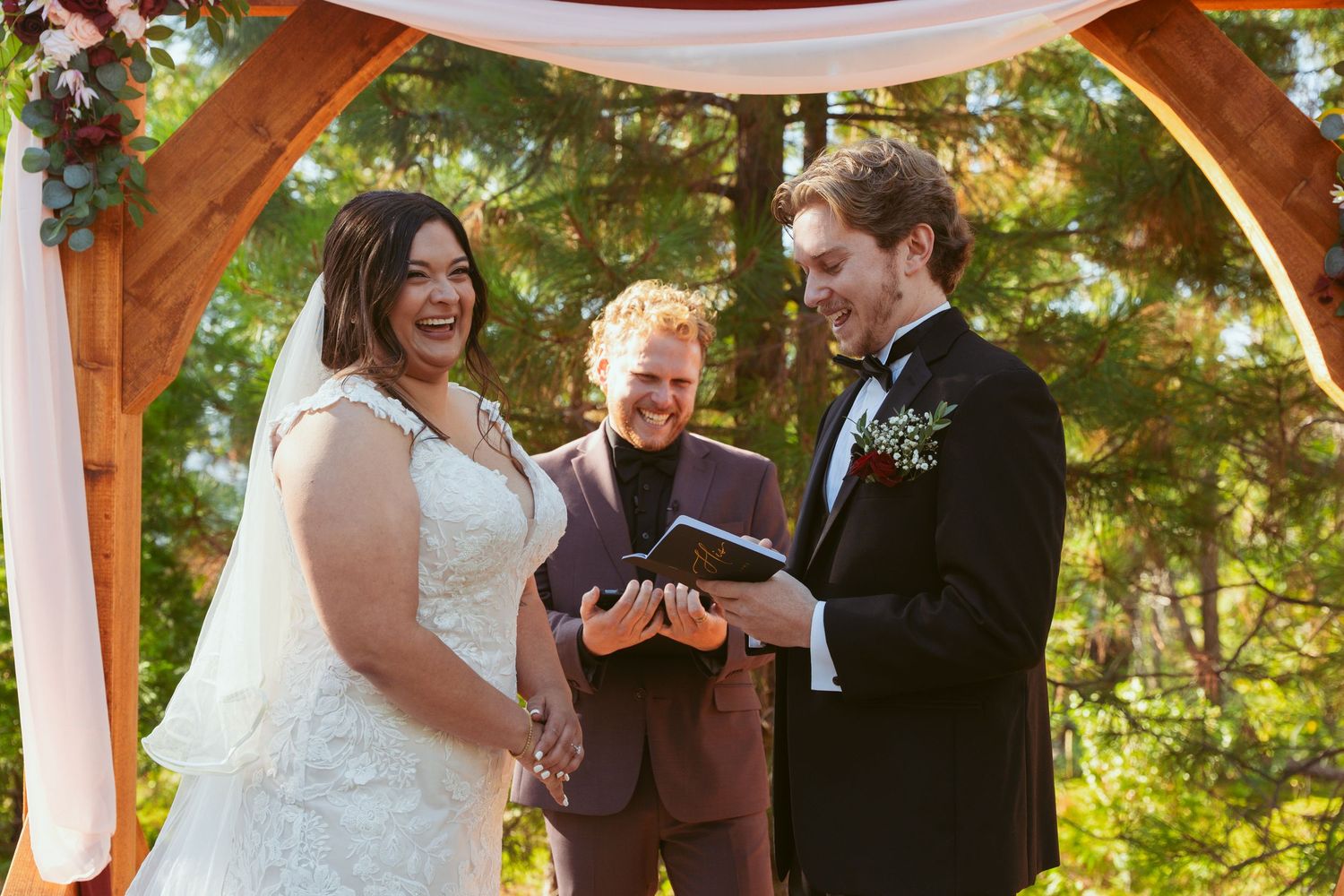 Intimate forest wedding ceremony with wooden pergola and white fabric decor.