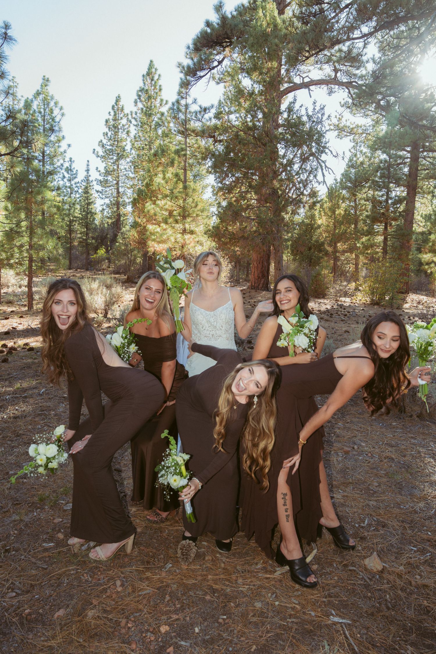 Bridesmaids in black dresses pose playfully for photos in a forest setting.