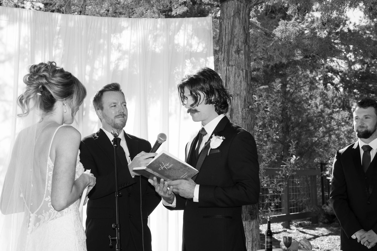 Wedding ceremony participants gather under white drapes and trees during an outdoor forest ceremony.