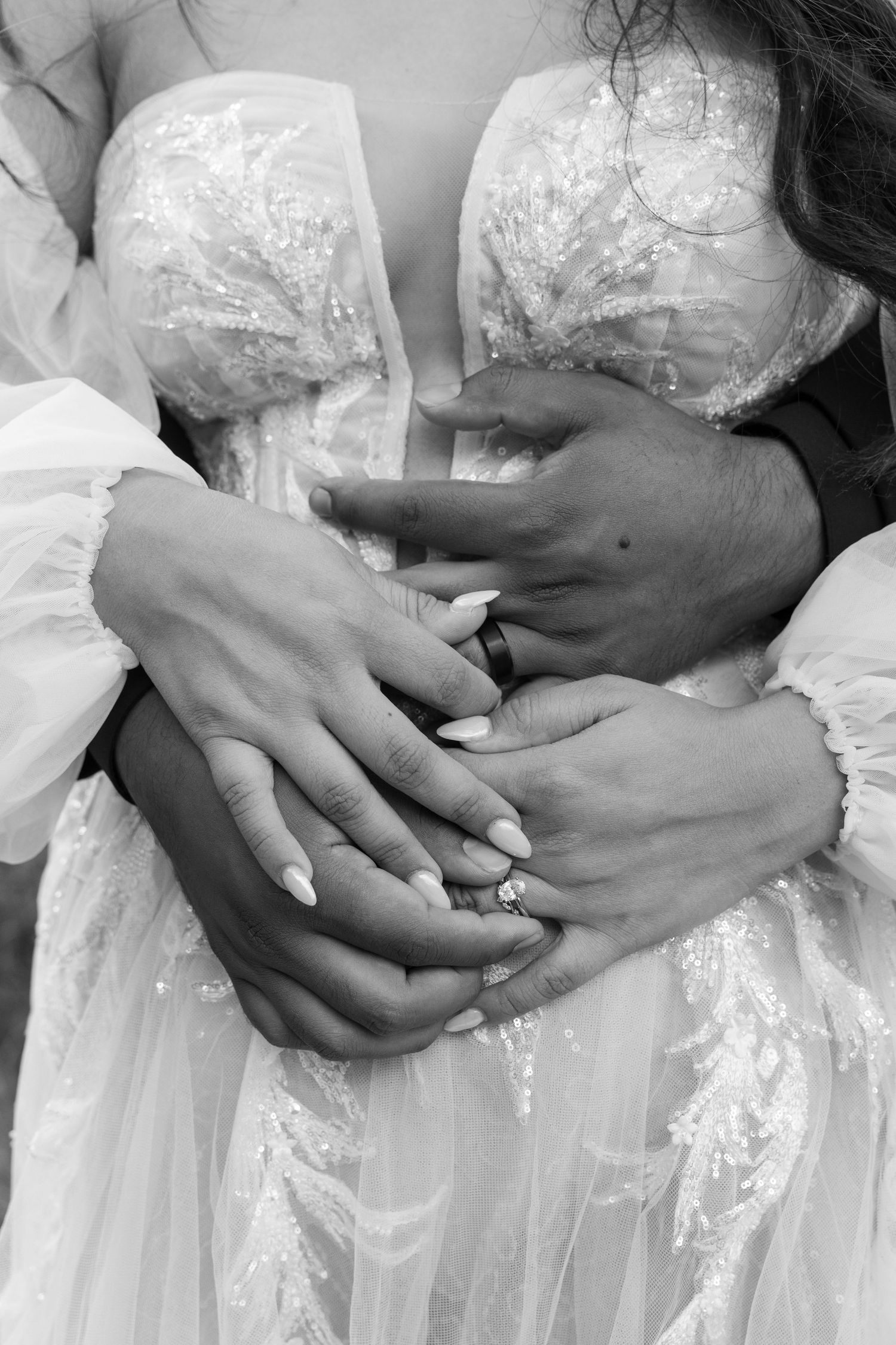 Black and white close-up of hands displaying wedding rings against delicate lace dress.