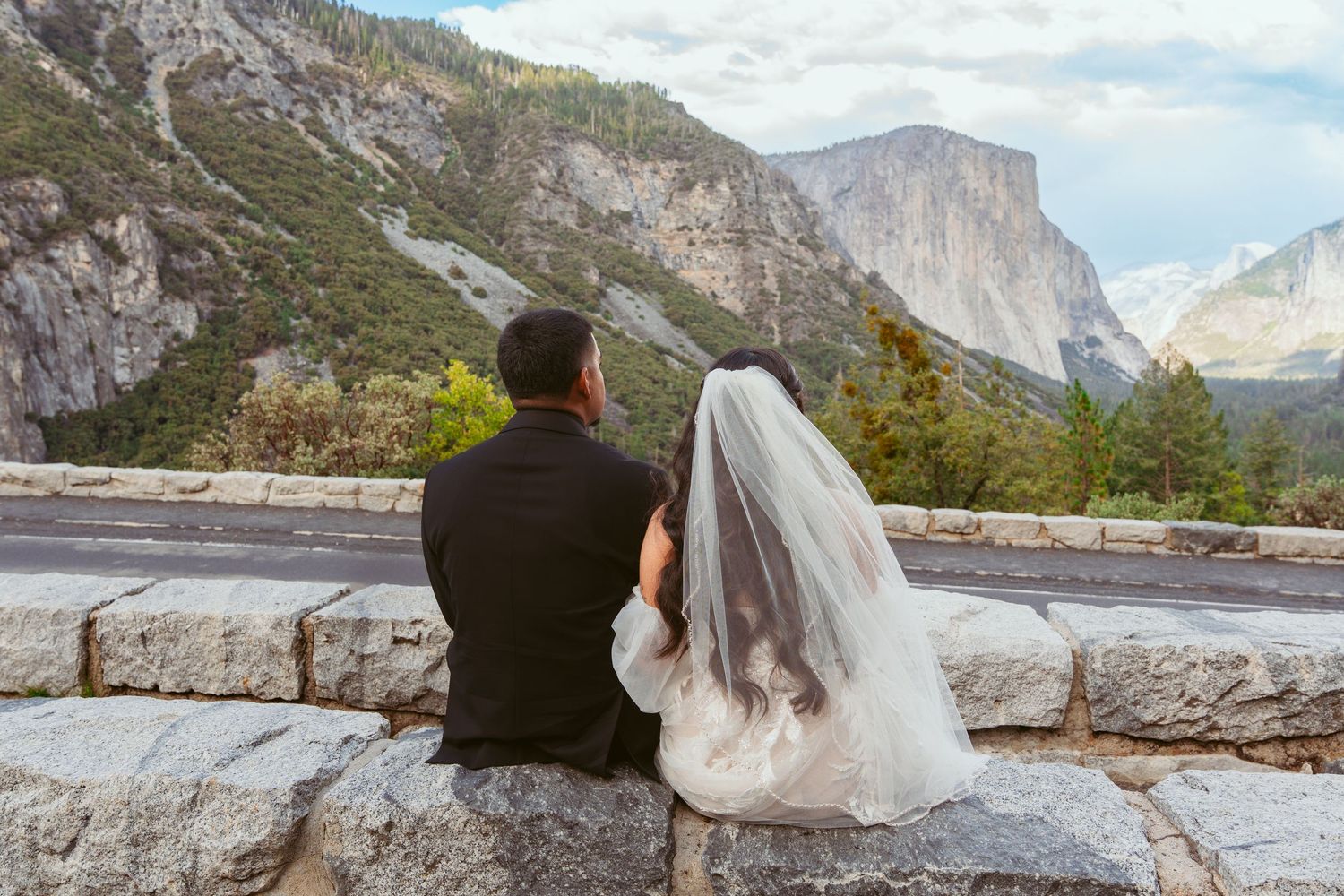 Distant shot of newlyweds sitting together on a stone wall overlooking Yosemite Valley.