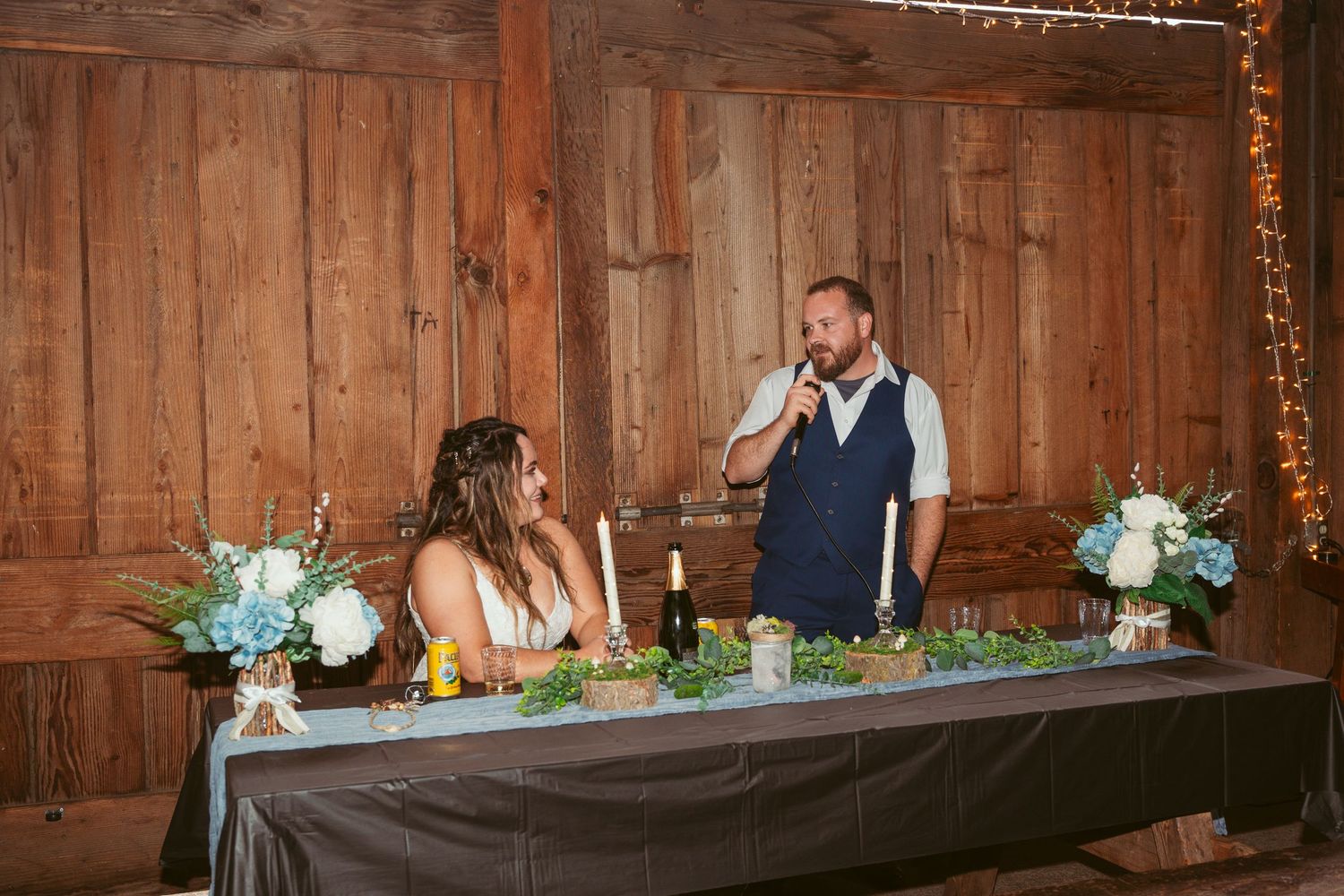 Wedding party seated at a long table inside a wooden barn with decorative lighting and floral arrangements.