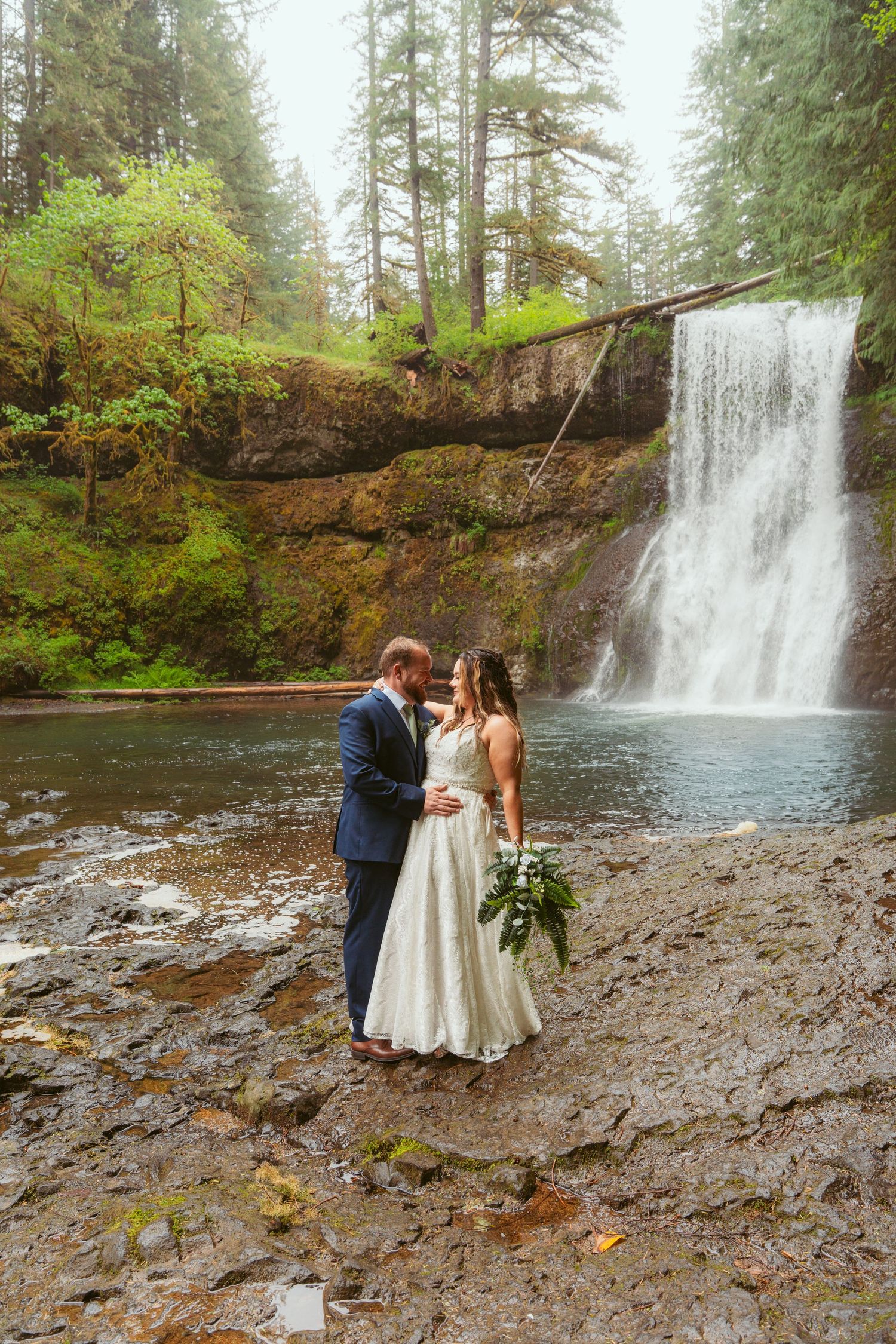 Newlyweds share tender moments by a forest waterfall during their wedding photo session.
