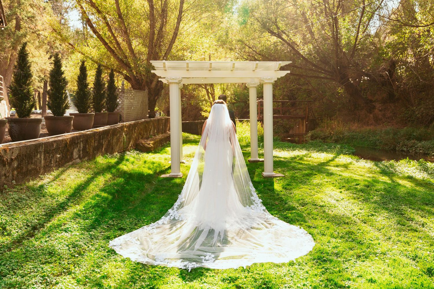 A bride in a flowing white dress stands in golden sunlight beneath a garden pergola.