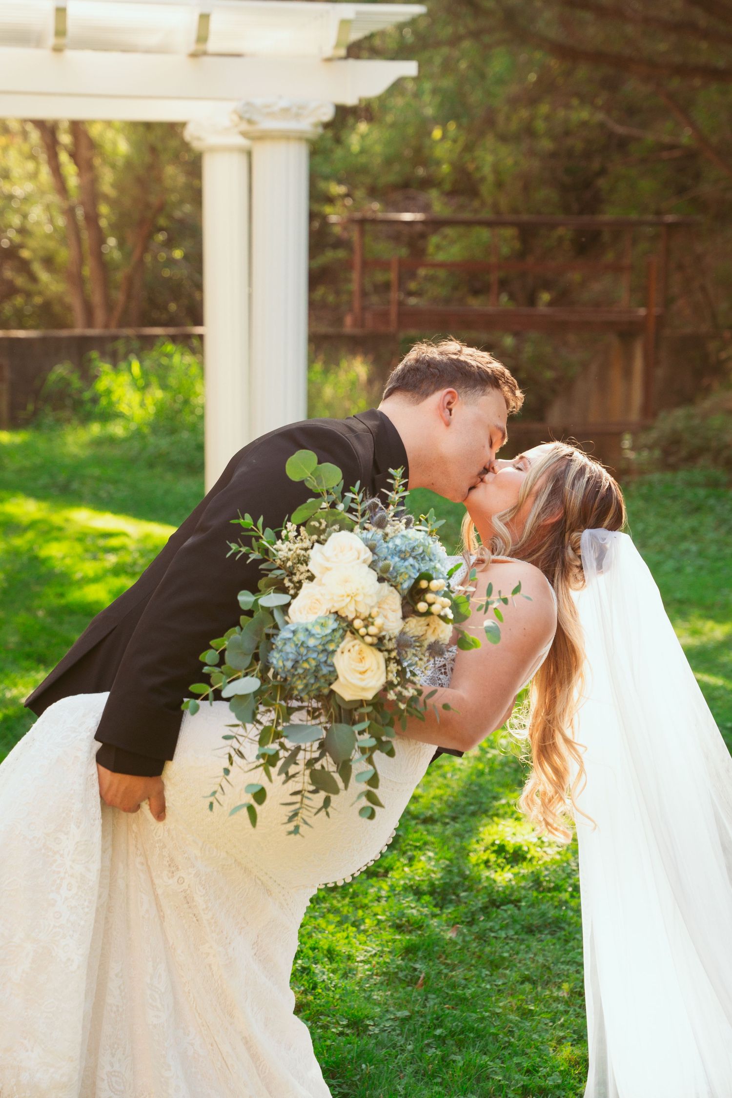 A romantic wedding couple shares tender kisses in a garden setting with white floral bouquets.