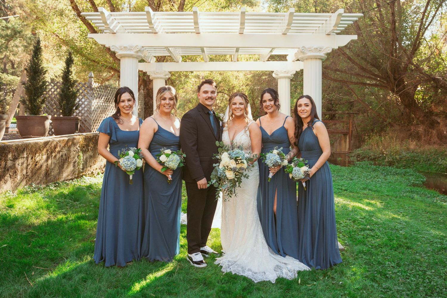 Bridal party in slate blue dresses poses outdoors under a white pergola on a sunny autumn day.