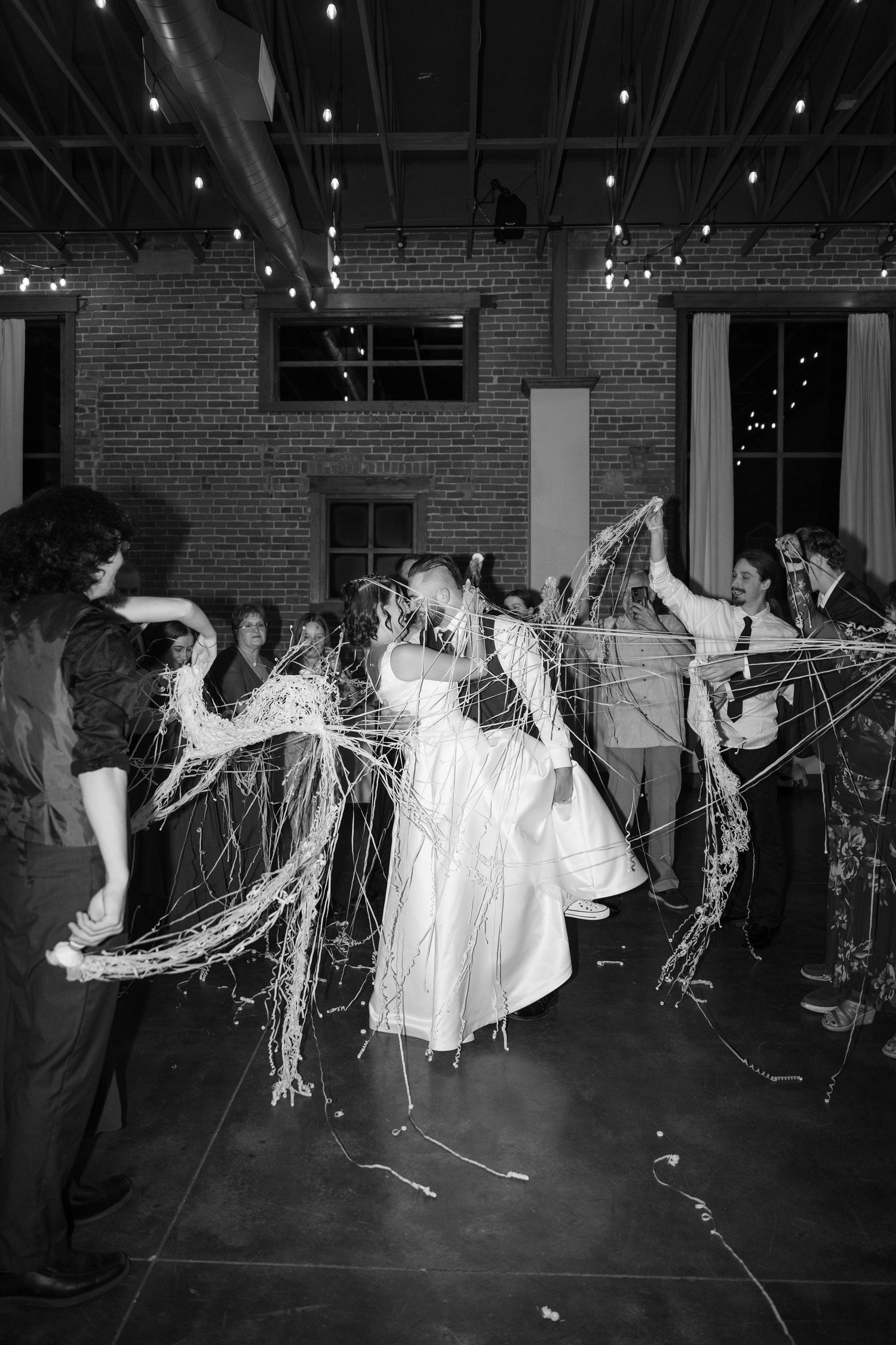 Black and white photo of party guests throwing streamers in an industrial-style venue with exposed brick.