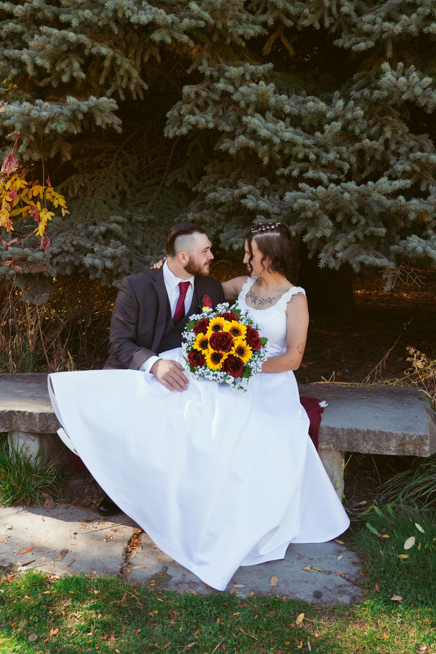 Newlyweds sitting on stone bench surrounded by evergreen trees in autumn setting.