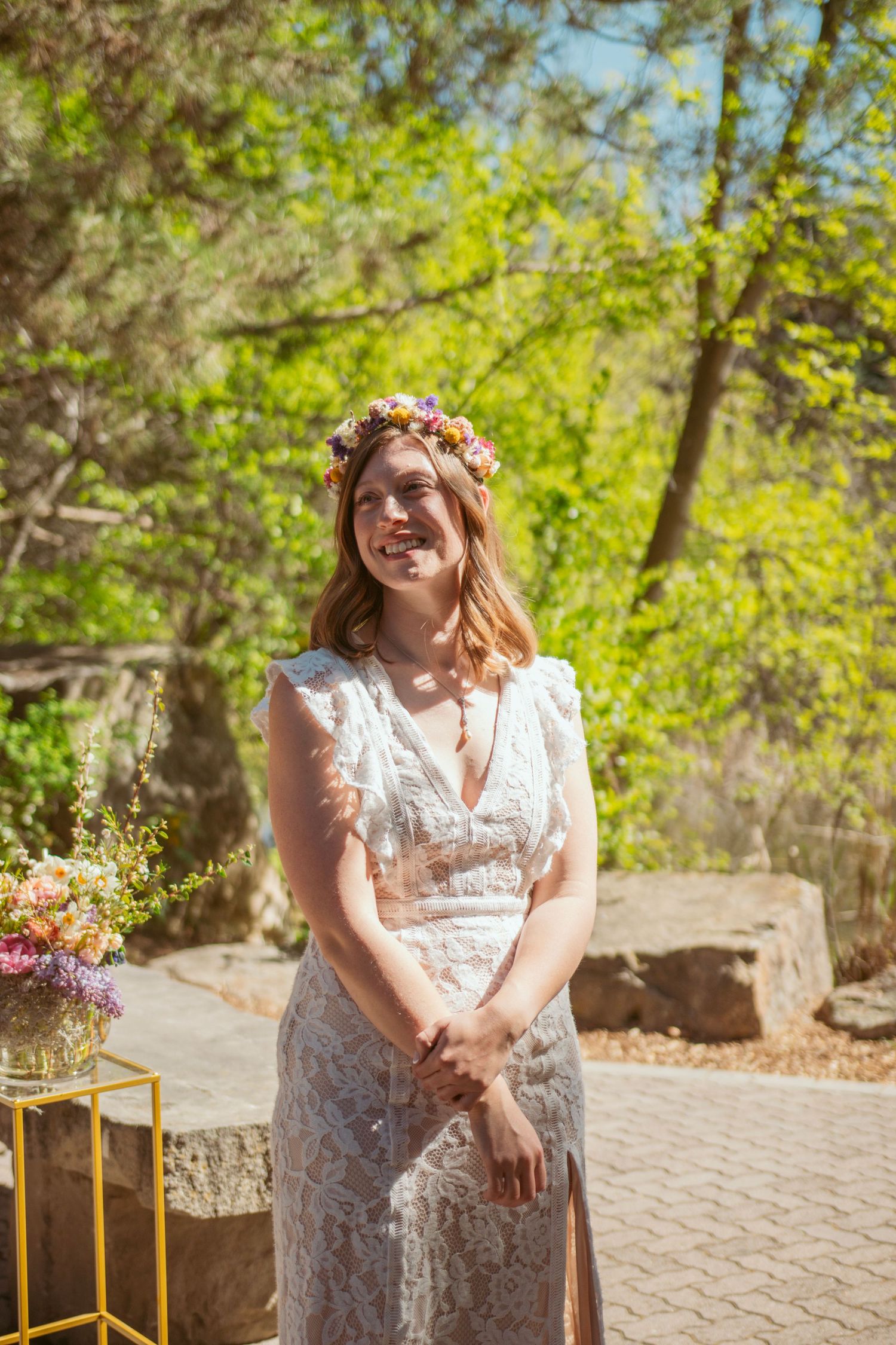 A bride in a lace wedding dress and floral crown stands outdoors in a garden setting with natural sunlight.