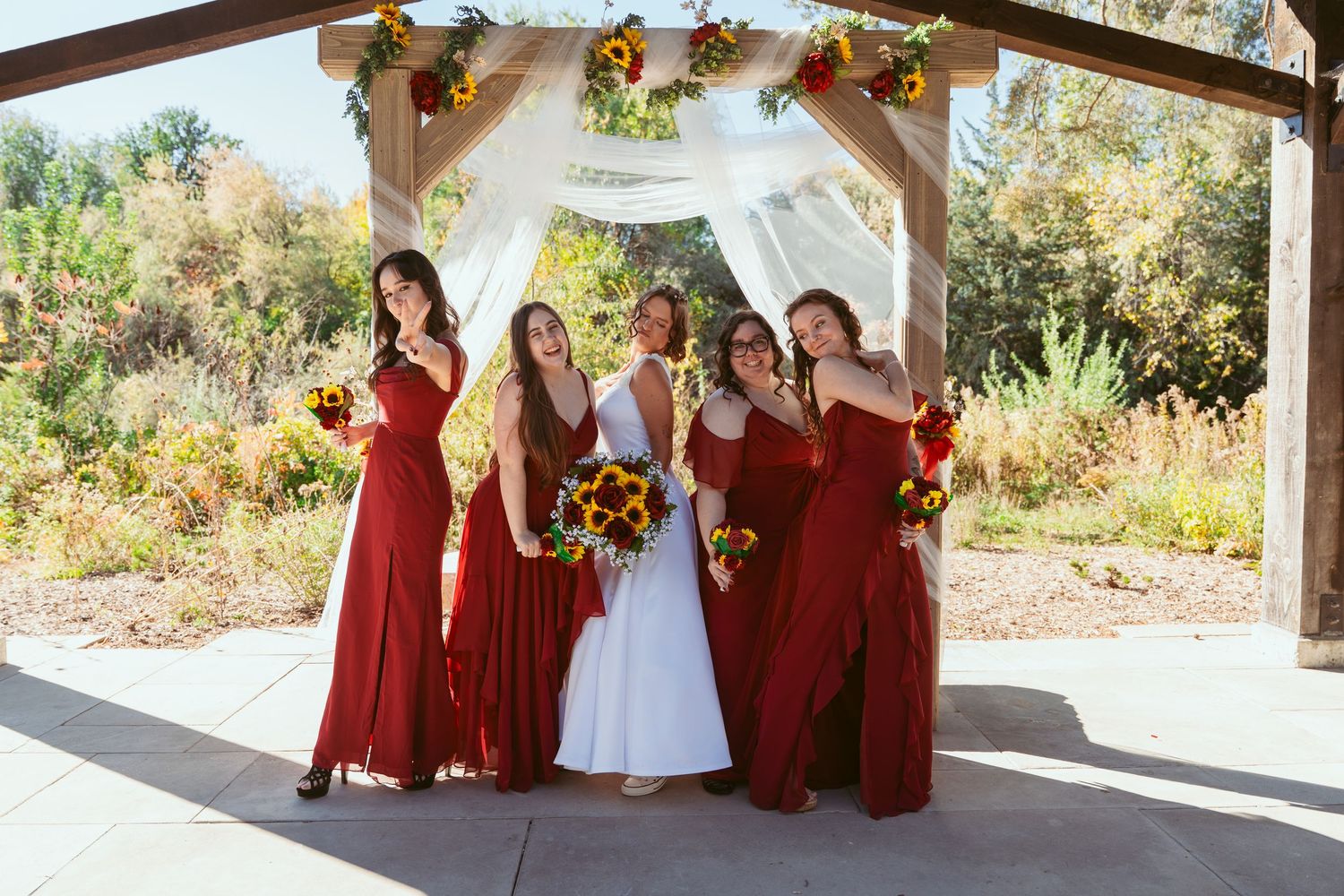 Wedding party poses in burgundy dresses and white gown under outdoor pavilion with sunflowers and roses.