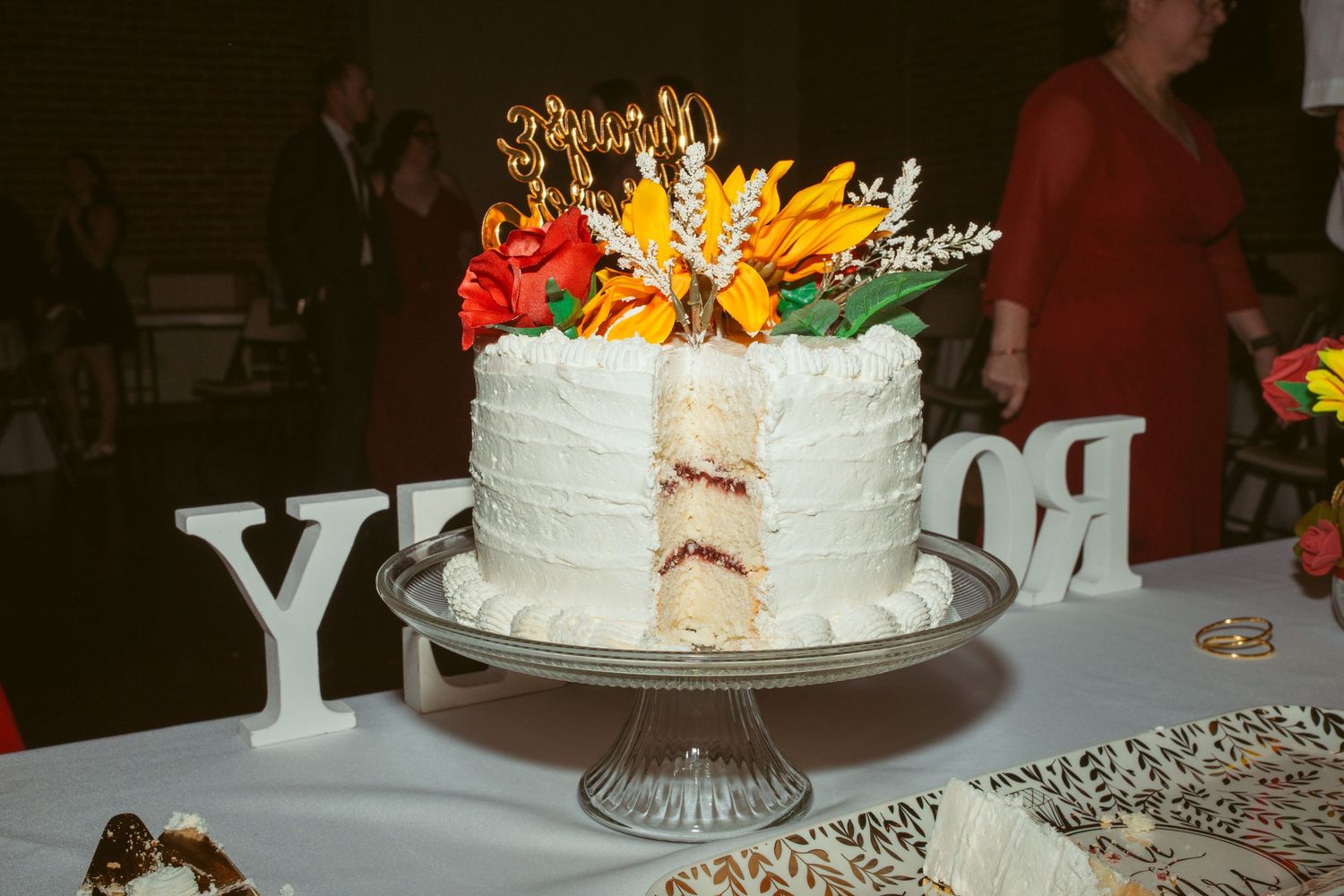 White wedding cake with colorful floral decorations displayed on a cake stand with decorative letters.