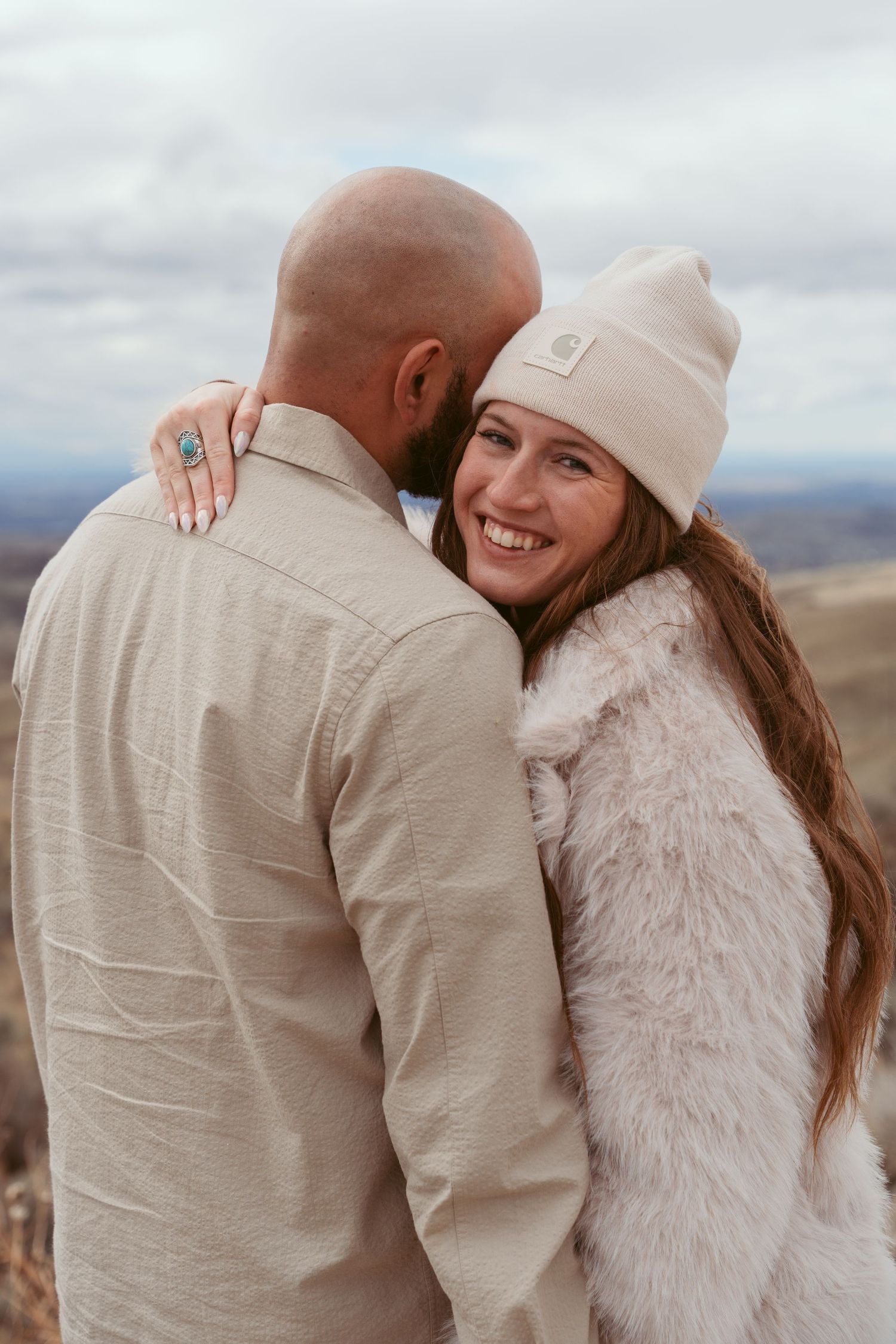 Couple embracing outdoors wearing beige and white clothing with mountains in background.