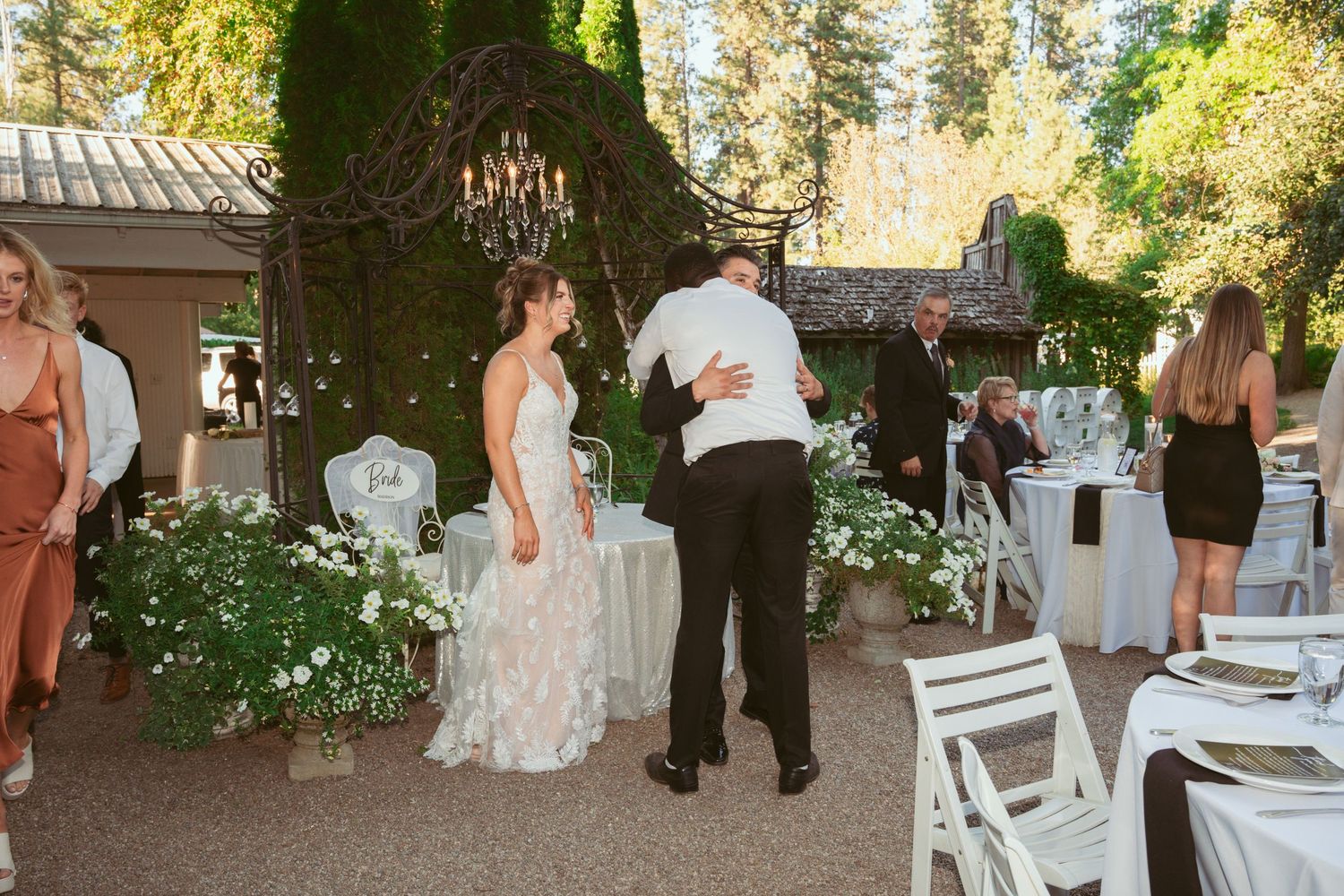 Outdoor wedding reception setup with white chairs, floral decorations and tall trees in the background.
