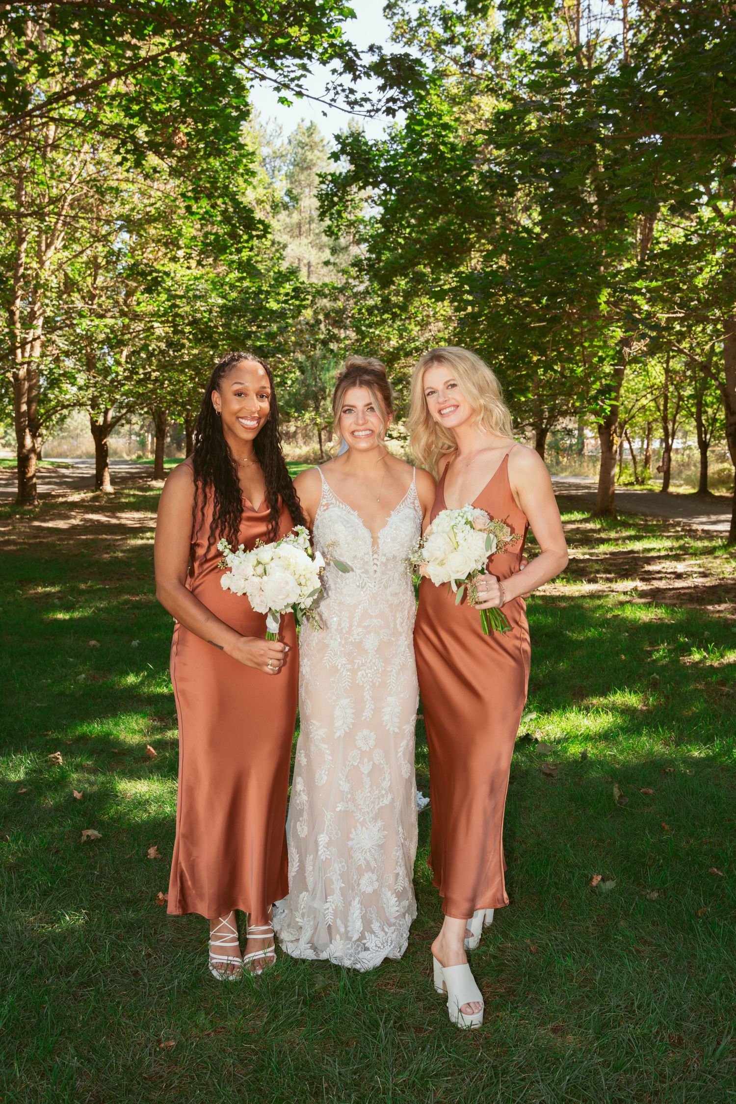 Wedding party in rust-colored bridesmaid dresses and lace bridal gown poses outdoors in natural setting.