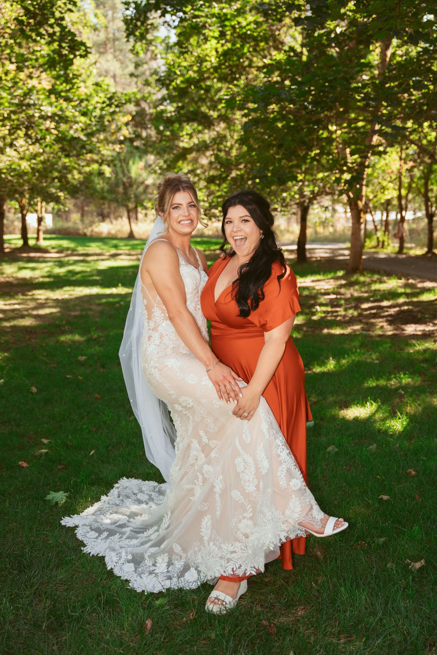 Wedding party members share joyful moments on grass under trees.