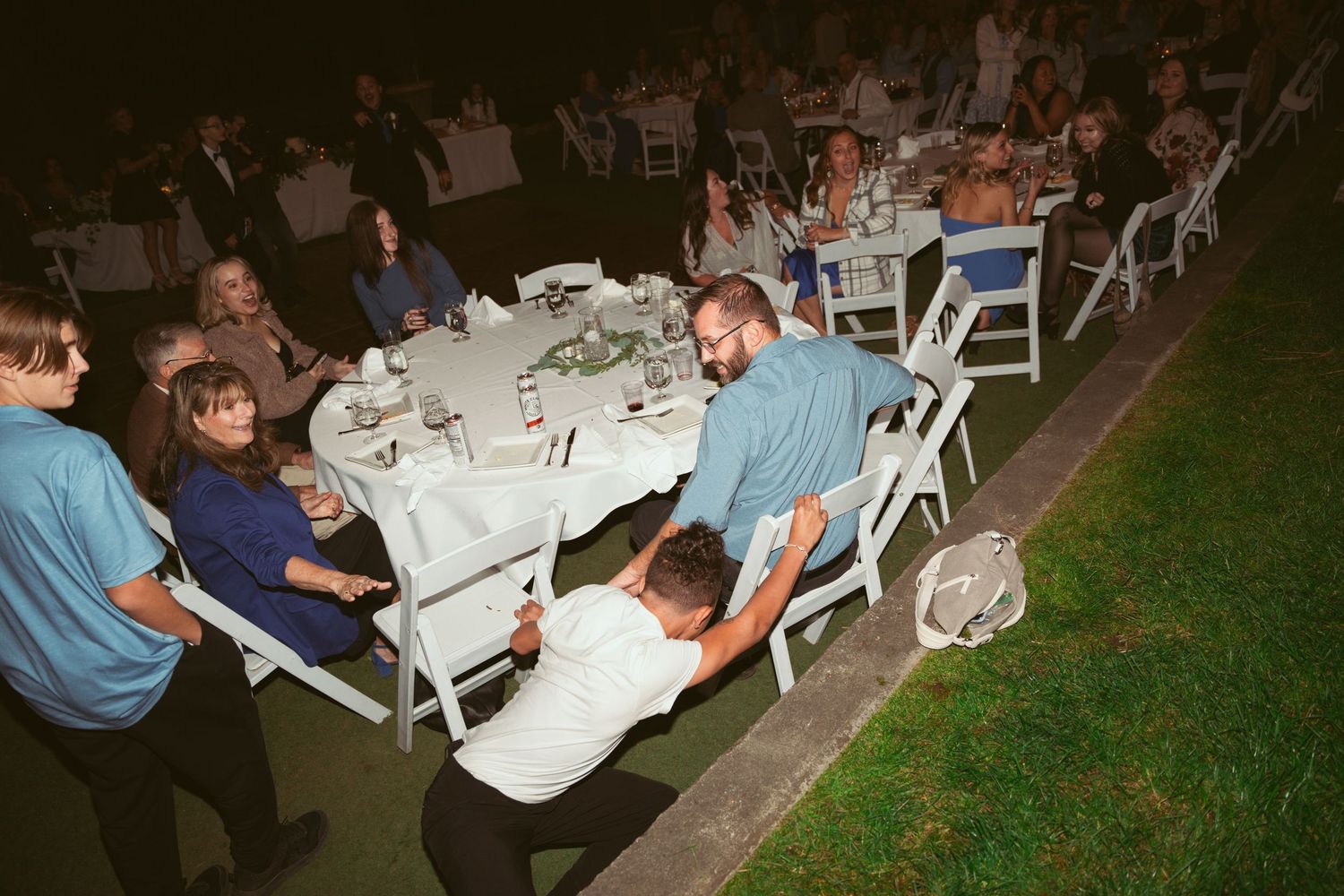 Wedding guests seated at outdoor reception tables with white chairs in evening lighting.