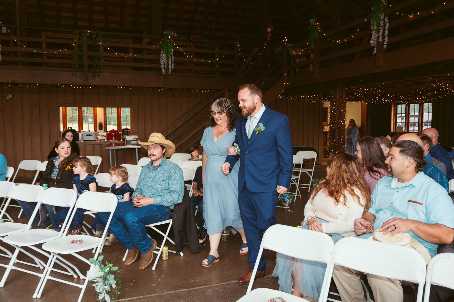 Wedding ceremony in rustic barn setting with guests seated on white folding chairs.
