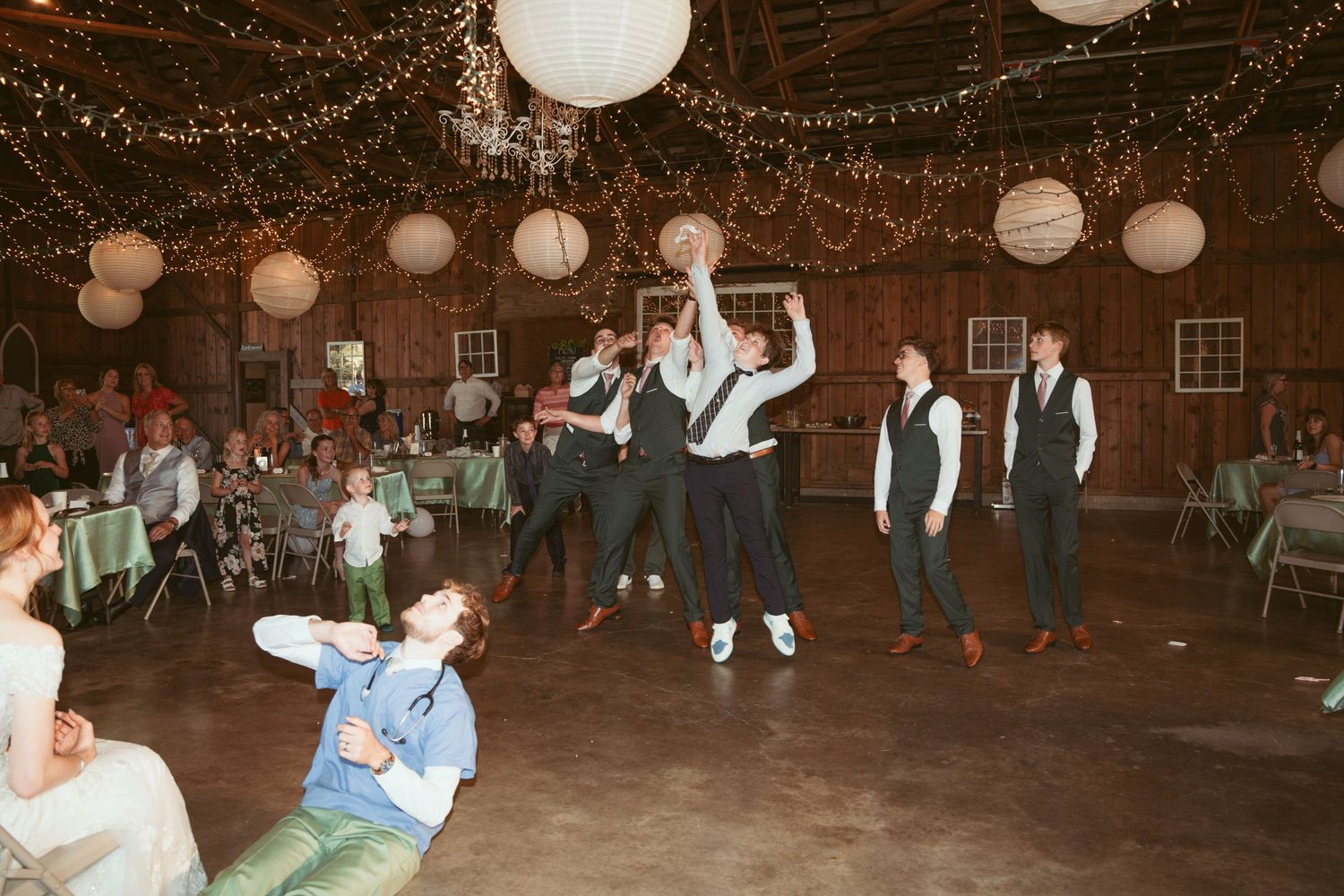 Wedding reception dance party in a rustic barn venue illuminated by string lights.