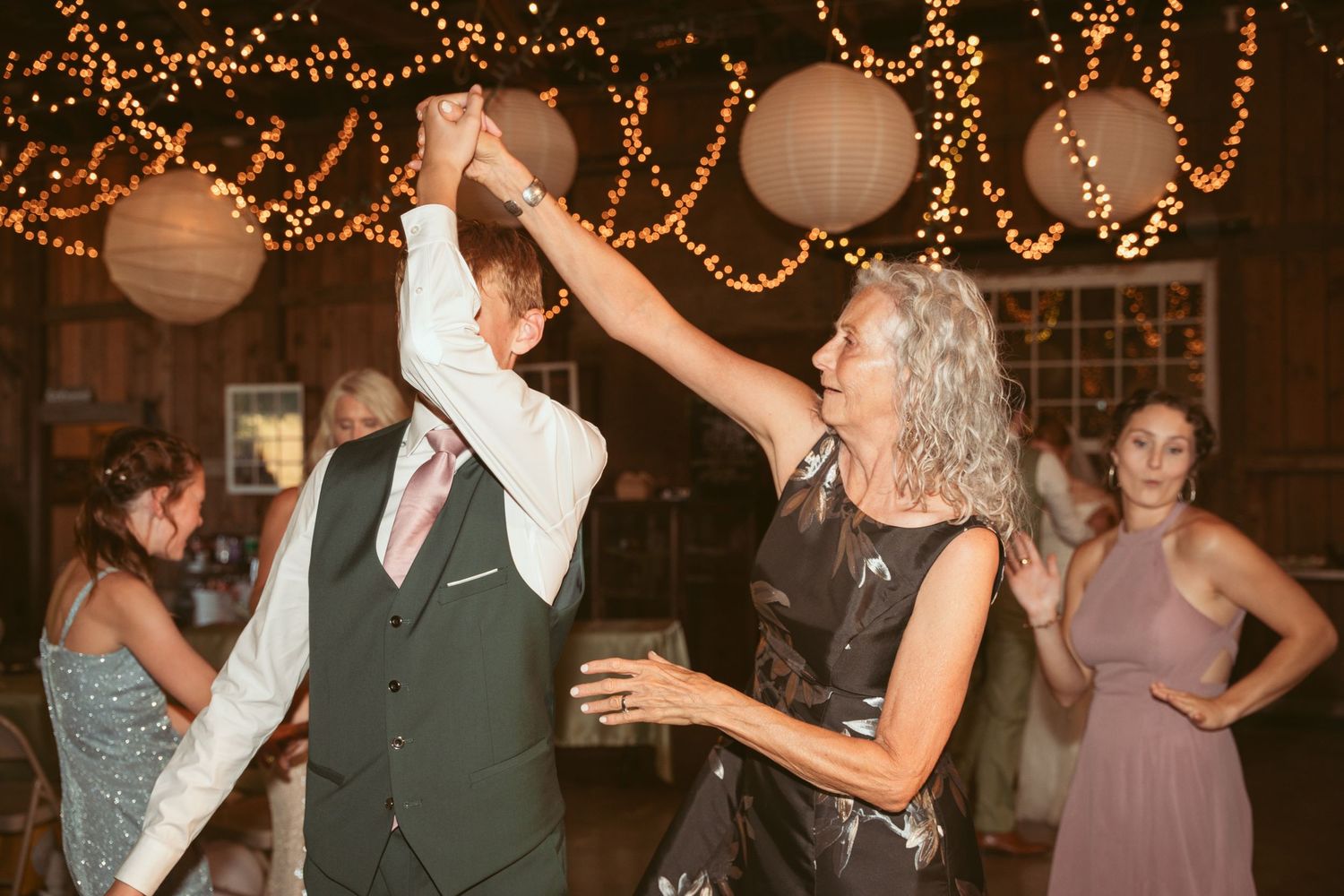 Party guests dancing energetically under decorative string lights and lanterns at an indoor venue.