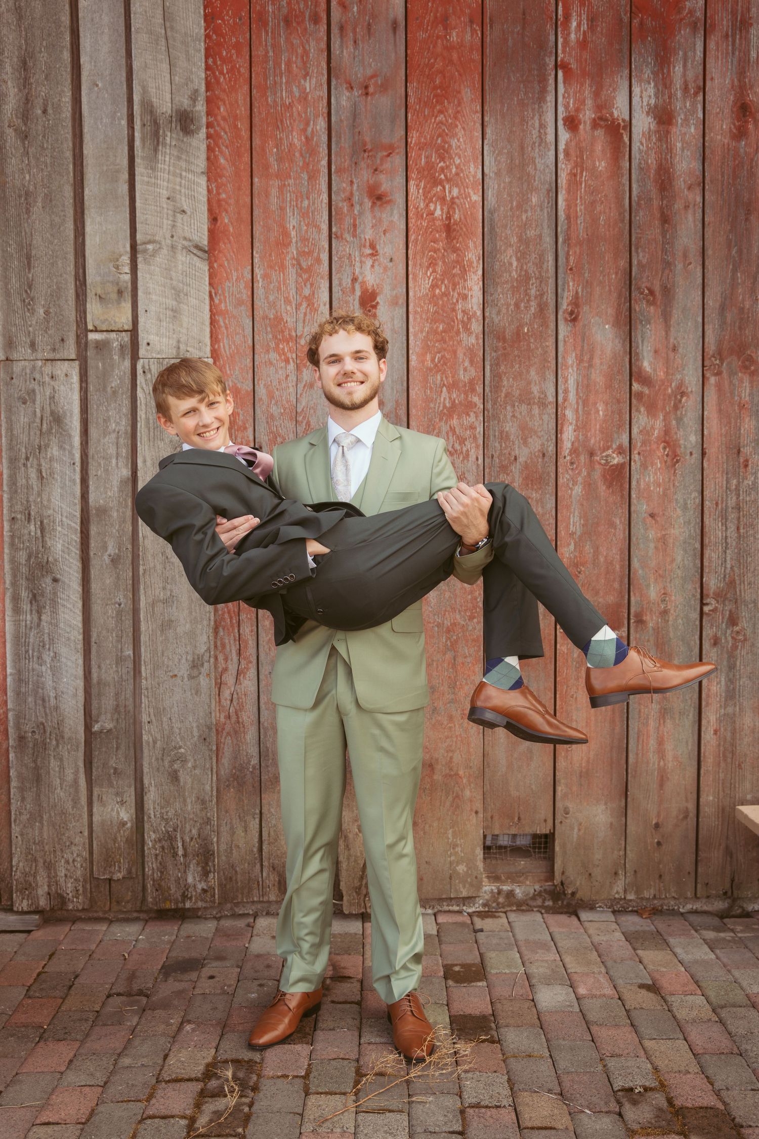 Playful wedding photo with one person carrying another in front of rustic barn setting.