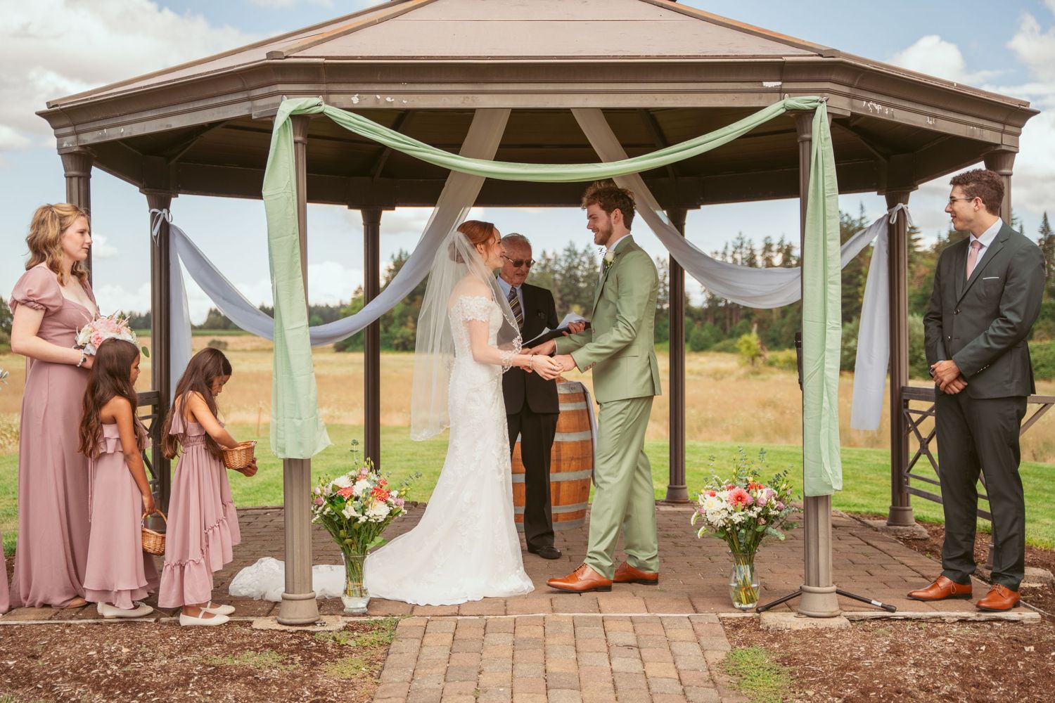 Wedding ceremony proceedings under rustic wooden gazebo with pastoral background.