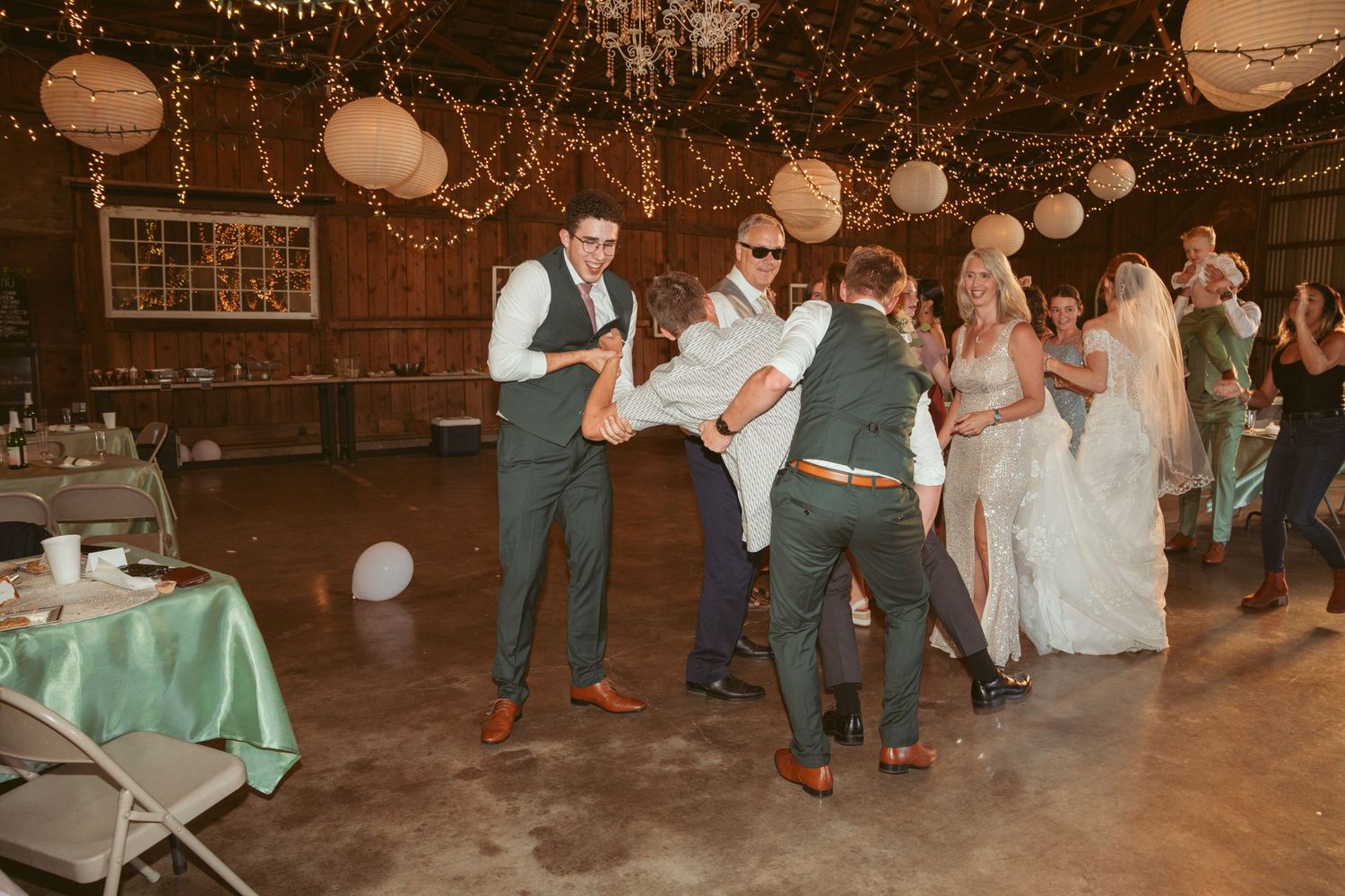 Wedding guests dance together under warm string lights and paper lanterns in a rustic barn venue.