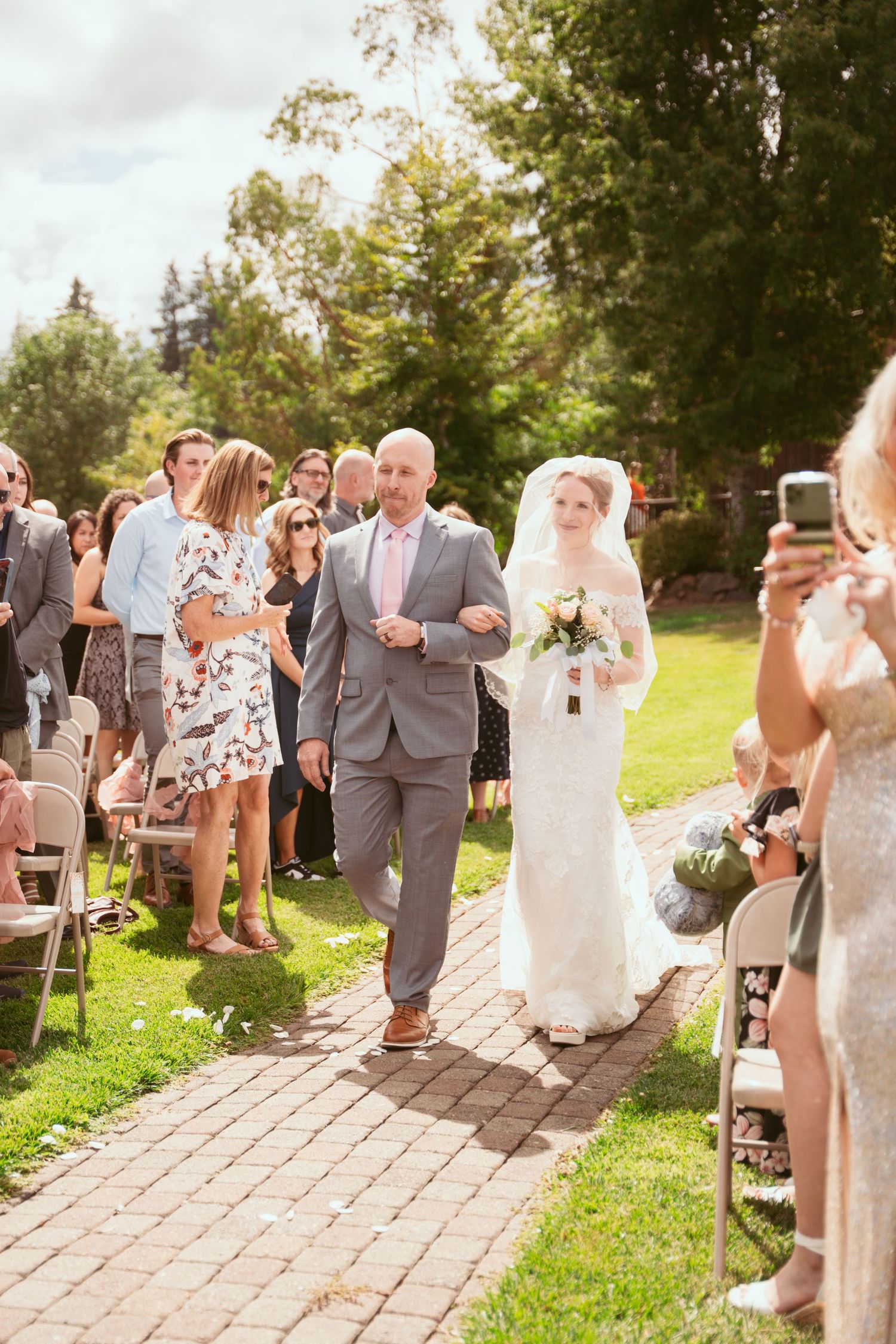 Wedding processional on brick pathway with guests seated on folding chairs under sunny skies.