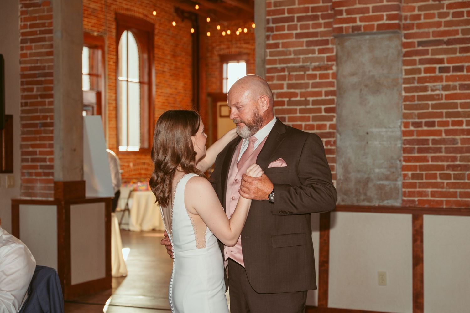 Couples share dances in an elegant brick-walled reception space with string lighting.