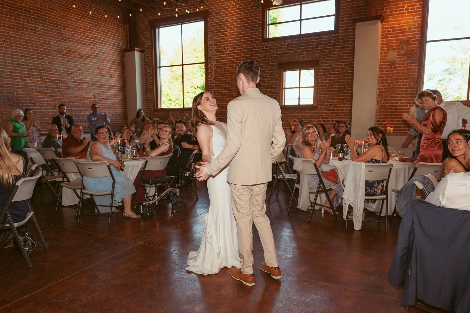 Wedding guests dance and celebrate on a rustic dance floor in a brick-walled venue with string lights overhead.