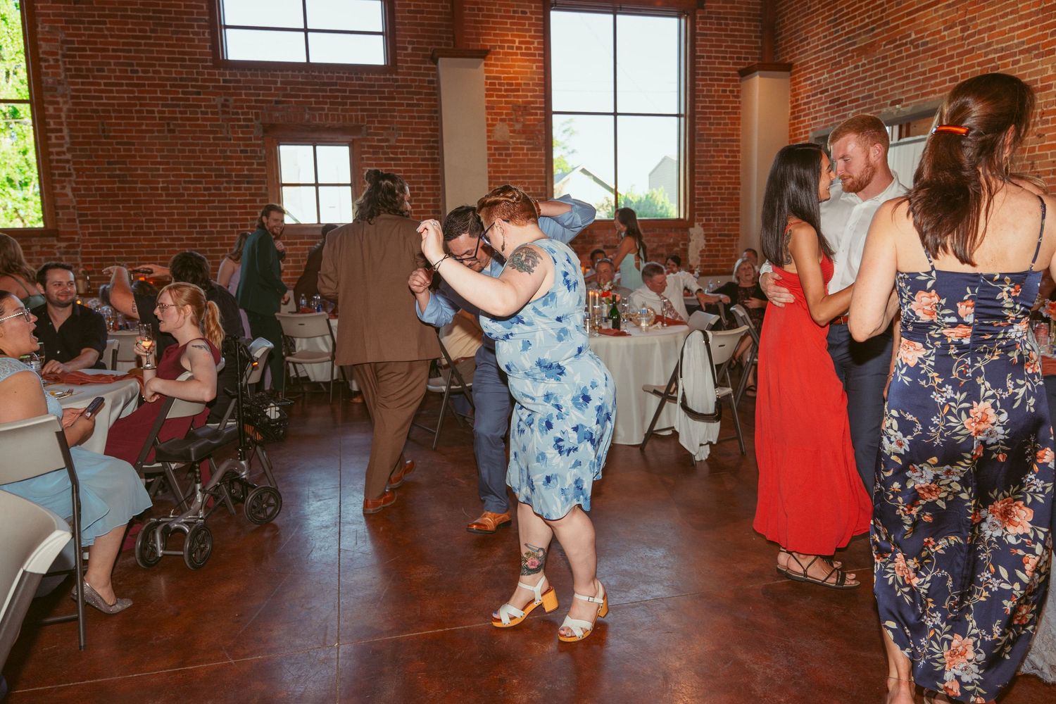 Wedding guests dancing together on a wooden dance floor in a rustic brick venue with string lights overhead.