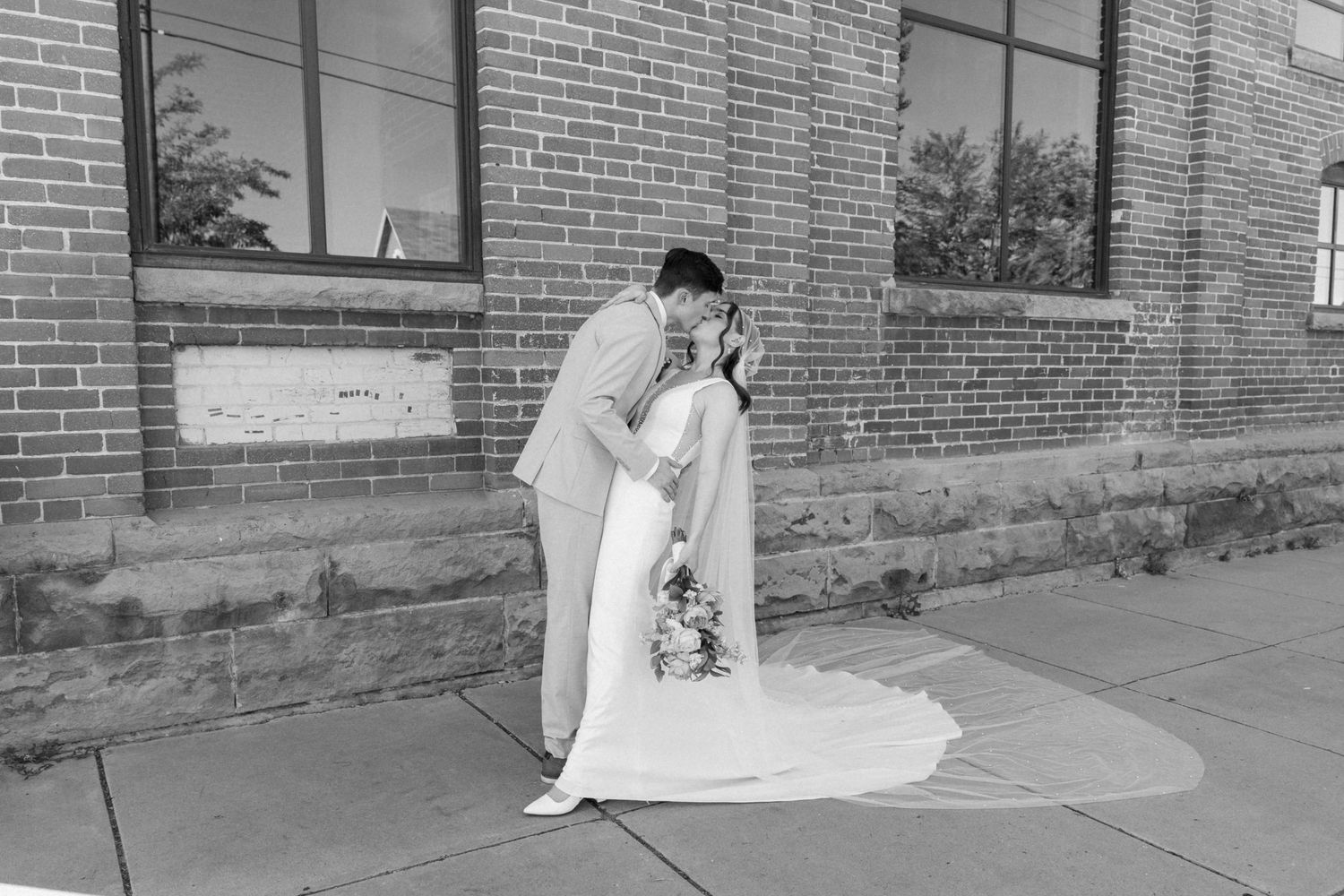 Wedding couple poses along brick building exterior in black and white photography.