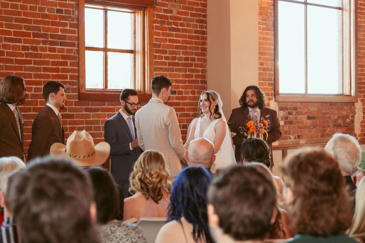 Wedding guests seated in rows watching ceremony in historic brick building with tall windows.