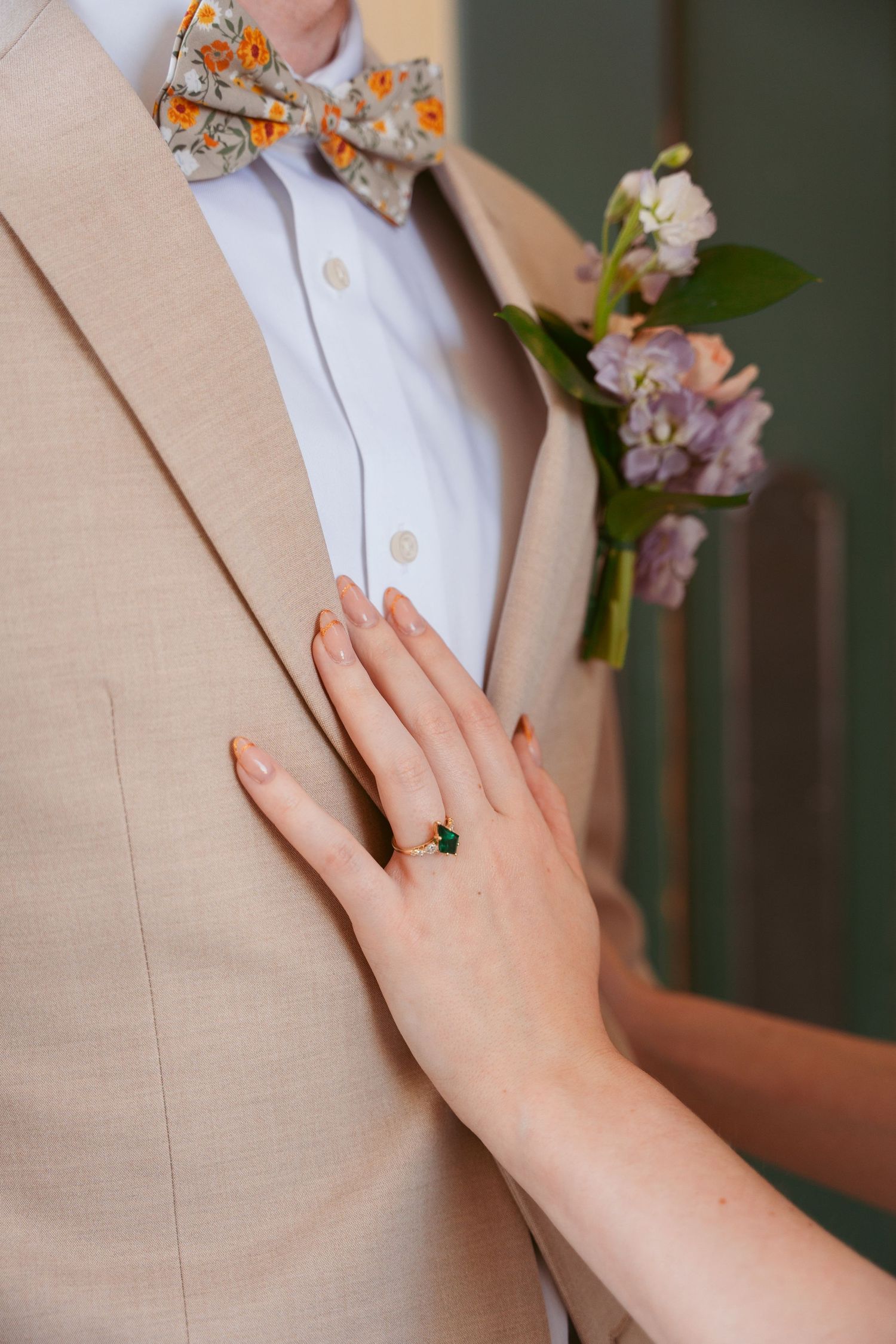 Close-up of engagement ring and floral boutonniere on beige suit jacket.