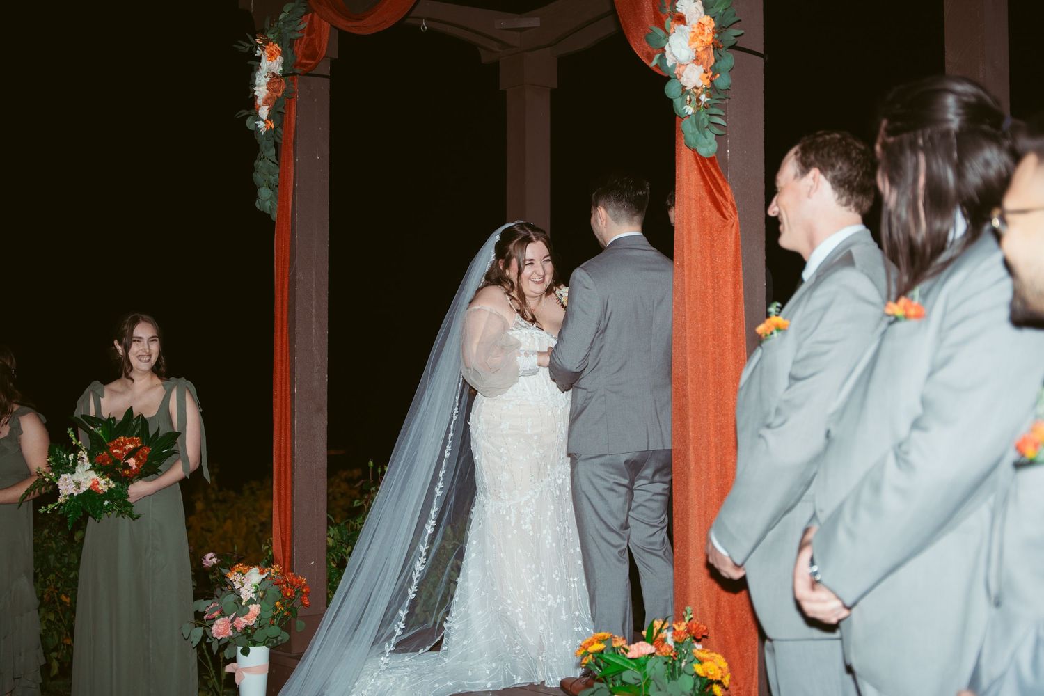 Evening wedding ceremony with bridesmaids in sage green dresses under a red-decorated arch.