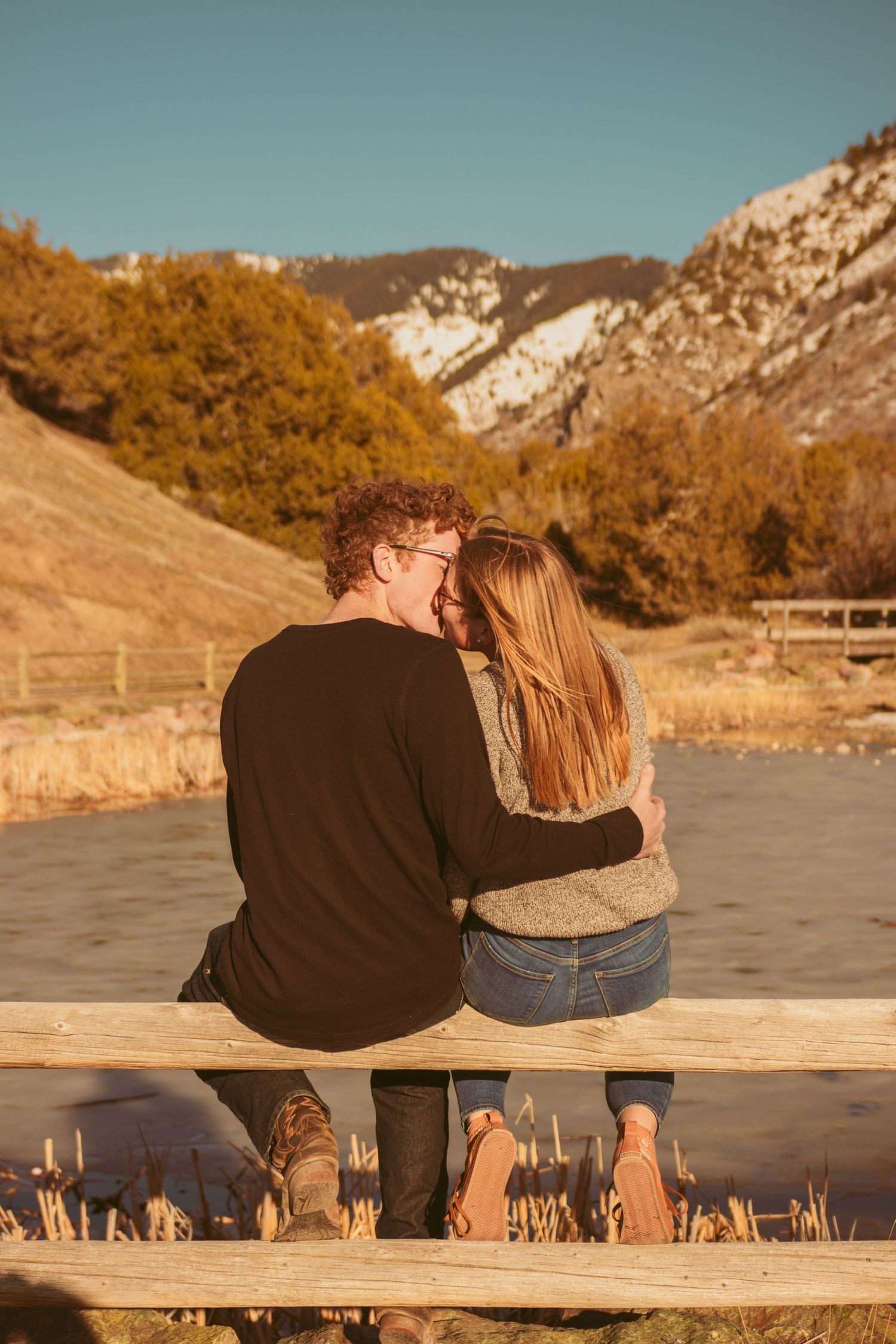 A couple shares an intimate moment while sitting on a wooden bench against a mountainous backdrop.