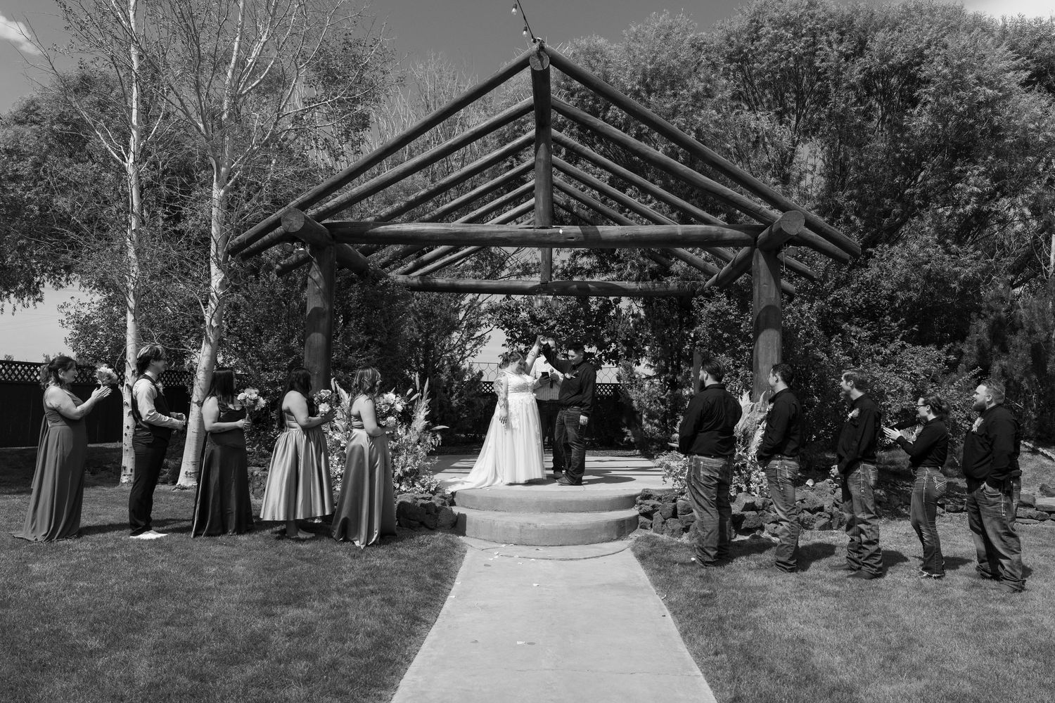 A black and white panoramic view of a wedding ceremony taking place under an outdoor wooden pavilion structure.