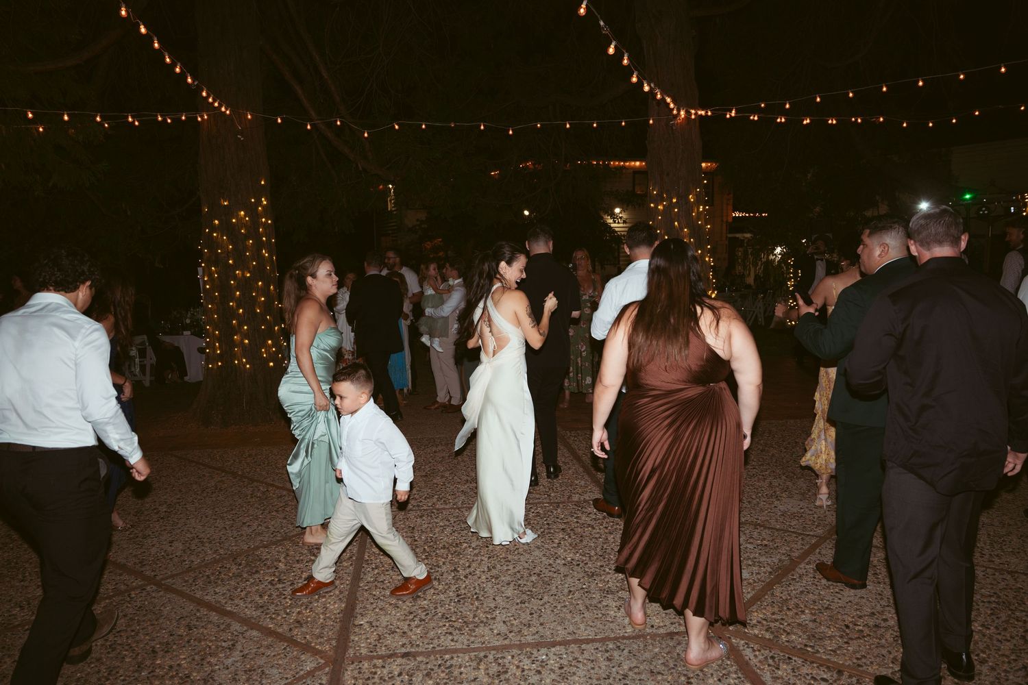 Wedding party dances beneath strands of twinkling lights at an outdoor evening celebration.