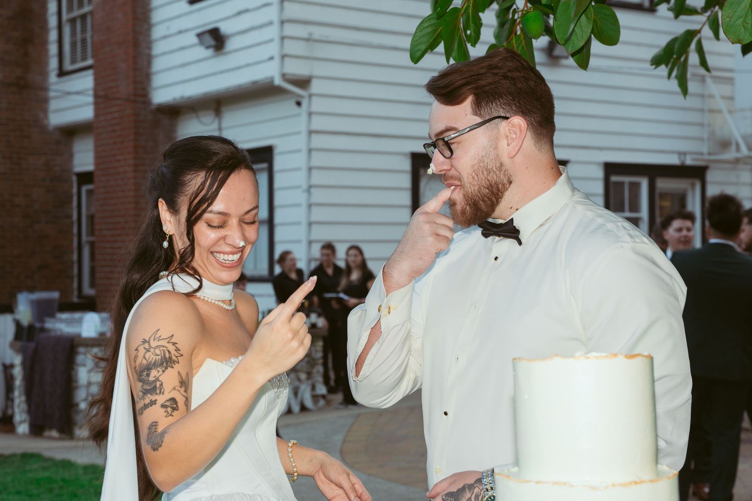 A couple in wedding attire shares cake during their reception outside a white building.