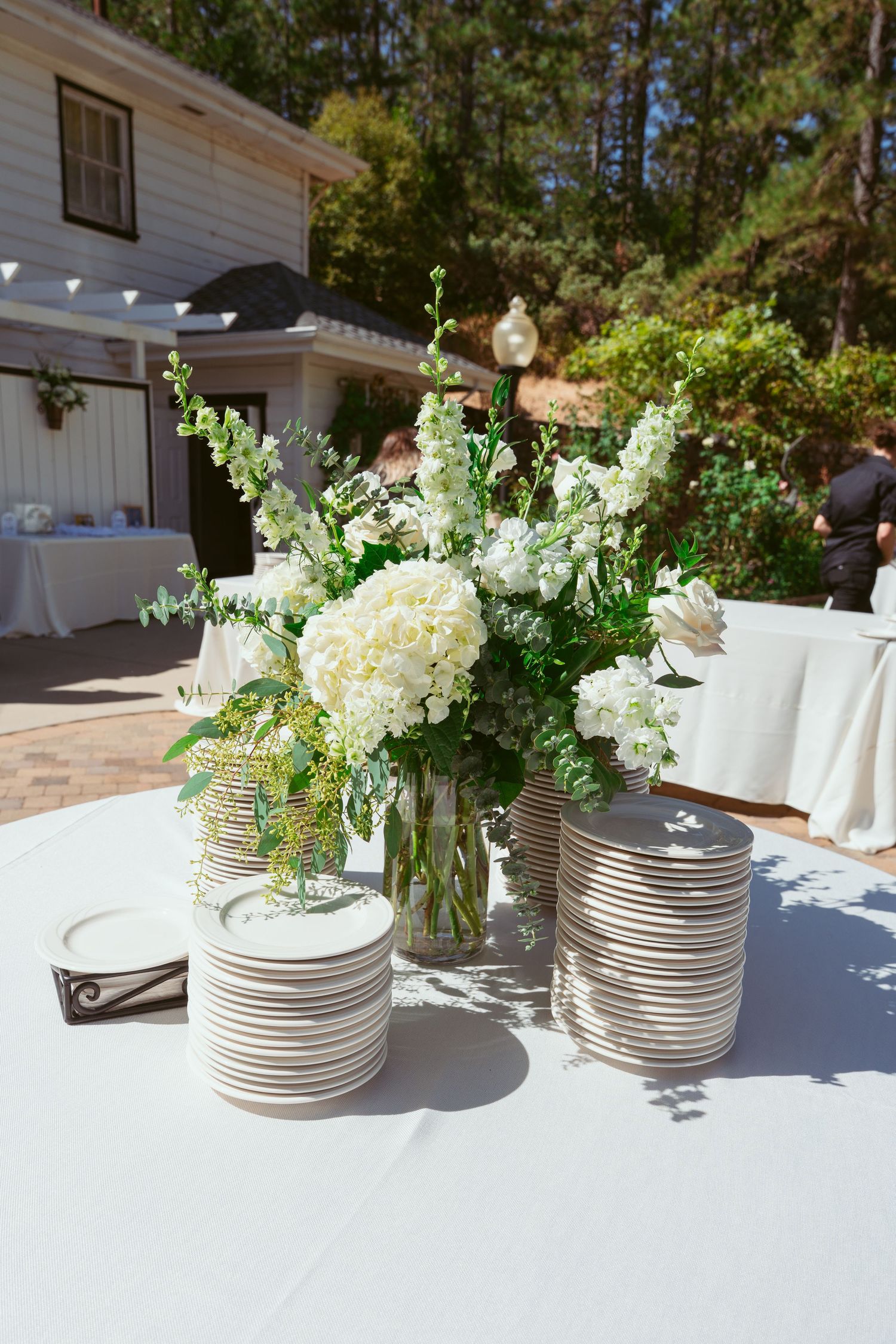 White floral centerpieces in silver tin vases arranged on a long table with white linens.